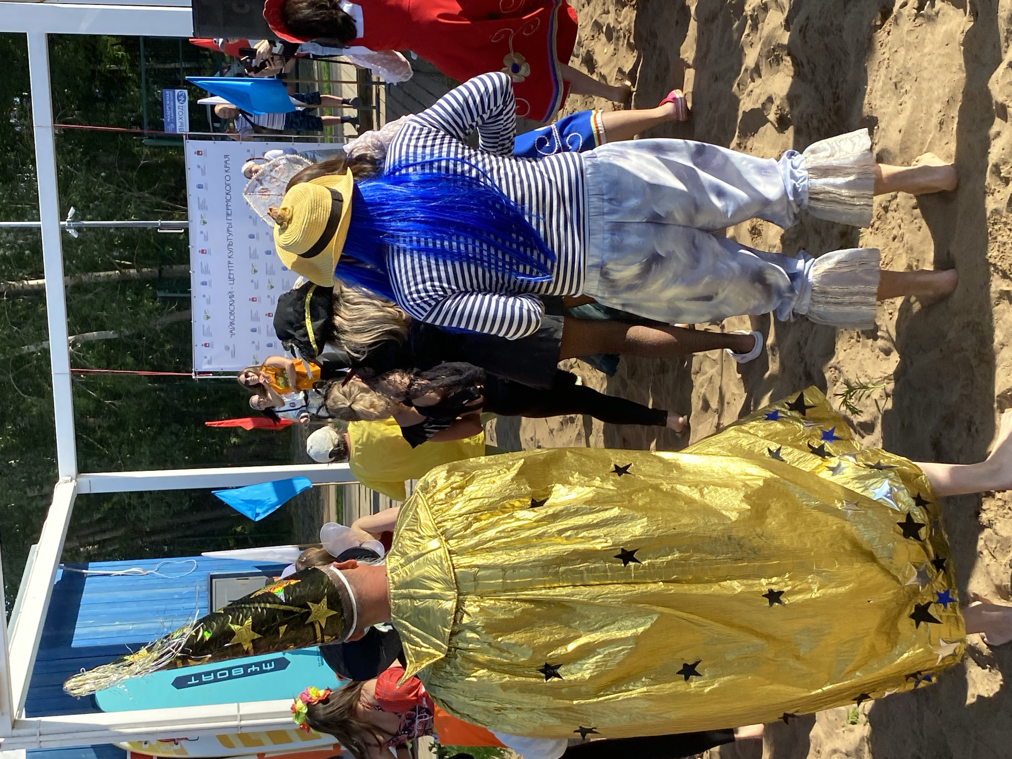In Chaikovsky, Russia, on a sunny day, a group of people in costumes are gathered on a sandy area with a blue building structure and trees in the background. The focus is on individuals in the foreground. One person wears a bright gold, star-spangled cape and a tall, pointed hat adorned with stars. Another person has bright blue hair and wears a striped long-sleeved shirt with light-colored, tie-dye pants that have ruffled bottoms. A woman in a red dress with floral accents is visible on the right. Other individuals in various costumes are partially visible, suggesting a festive event or gathering taking place outdoors. Flags, including a blue one, are planted in the sand. A large banner with Cyrillic text is displayed in the background.