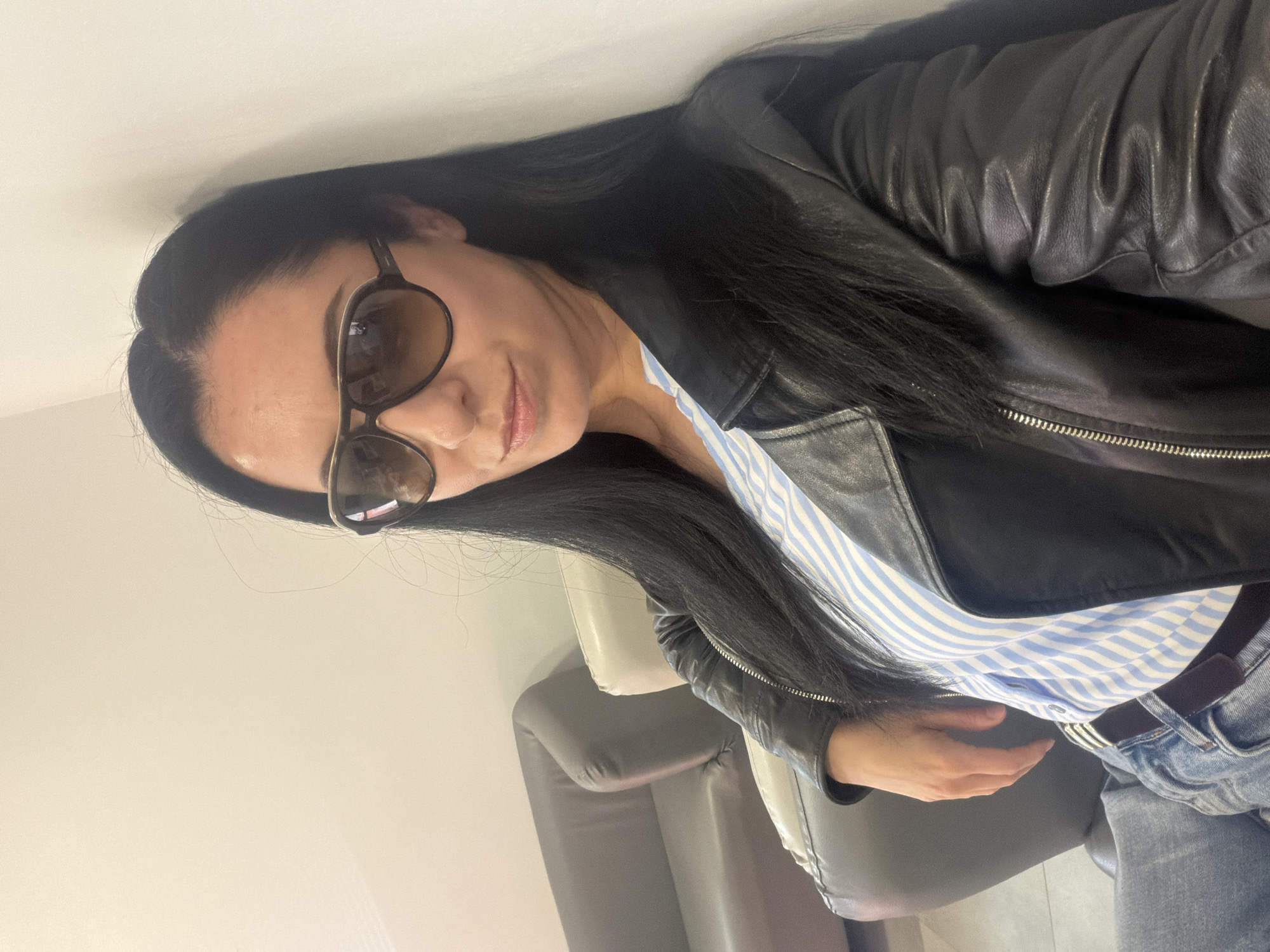 A woman with long, black hair and wearing sunglasses is the primary subject of the image. She is wearing a black leather jacket over a blue and white striped shirt. She is also wearing jeans with a brown belt. The woman appears to be indoors, near a white wall and next to a gray chair. The location of the image is in the city of Unknown, Unknown.