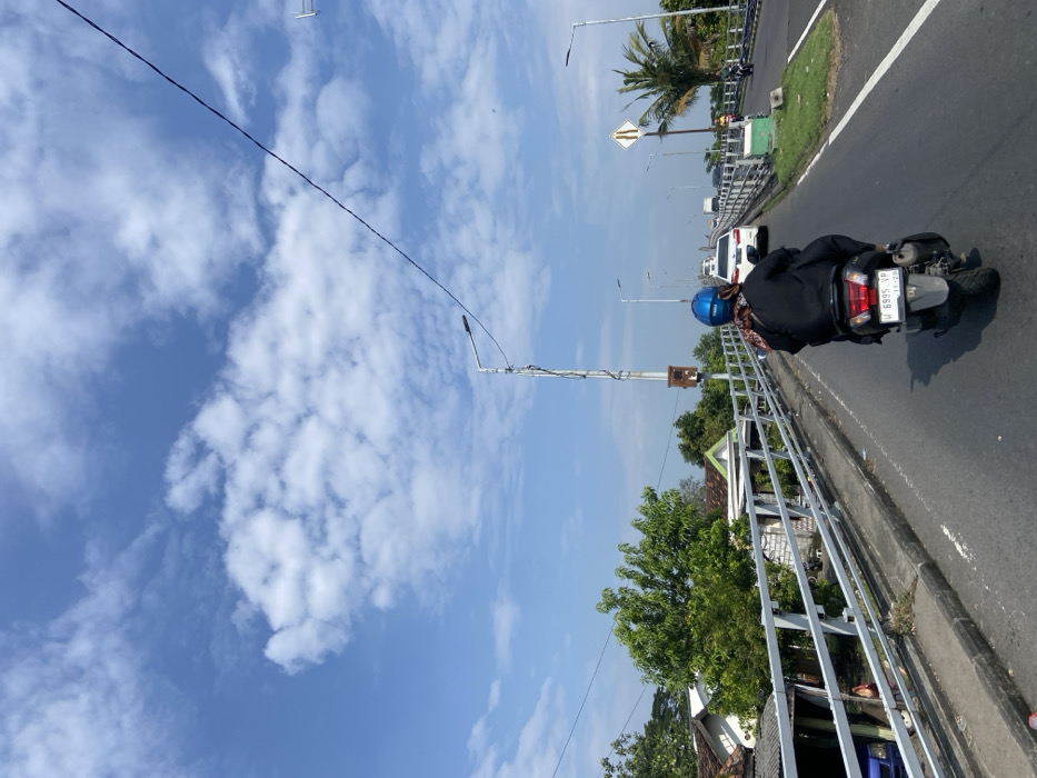 The image shows a person riding a motorcycle on a bridge in Kecamatan Tanggulangin, Indonesia. The person is wearing a blue helmet and is driving the motorcycle with a black license plate that reads "W 6995 VP." The bridge is surrounded by a silver metal guard rail. The road is asphalt and a white car is driving in the opposite direction. The sky is clear and blue with some white clouds. There is a street sign on the right side of the image. The bridge appears to be in a suburban or rural area with houses and trees visible in the background.