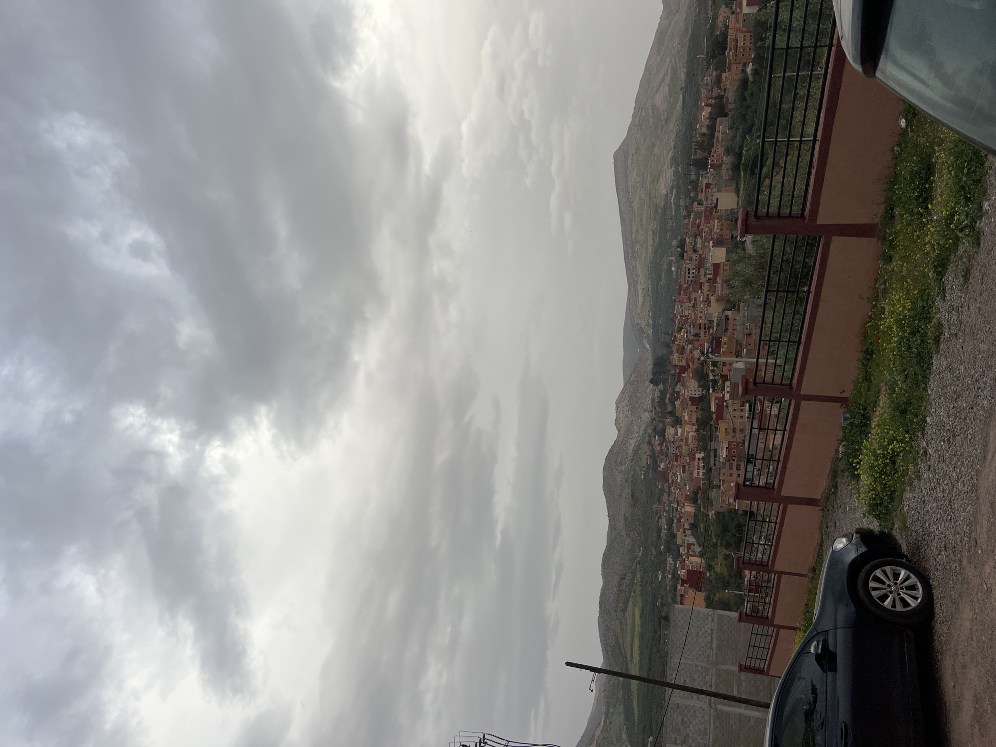 The image shows a cloudy landscape in Ouaouizert, Morocco. A town of closely packed, mostly orange-colored buildings nestles in the valley and climbs up a nearby mountain. A brown and black fence runs along the edge of a road, with green plants and yellow flowers growing next to it. Part of a black car is visible on the right side of the image. Power lines and poles are seen near the bottom of the image. The sky is overcast with grey clouds.