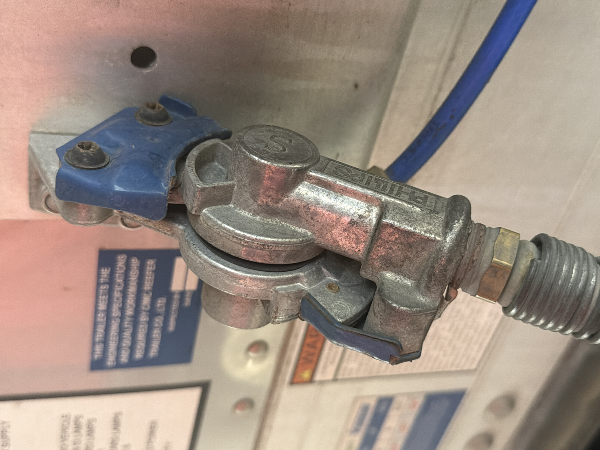The image shows a close-up of a metal coupling with a blue plastic cap, attached to a gray corrugated hose. The coupling has the word "PHILLIPS" embossed on it. In the background, there are labels on a light-colored surface. The scene appears to be an outdoor industrial or automotive setting, possibly at a truck stop or repair facility. The lighting suggests it is daytime. There is no indication of the city of Murfreesboro, United States of America, nor any people or specific actions.