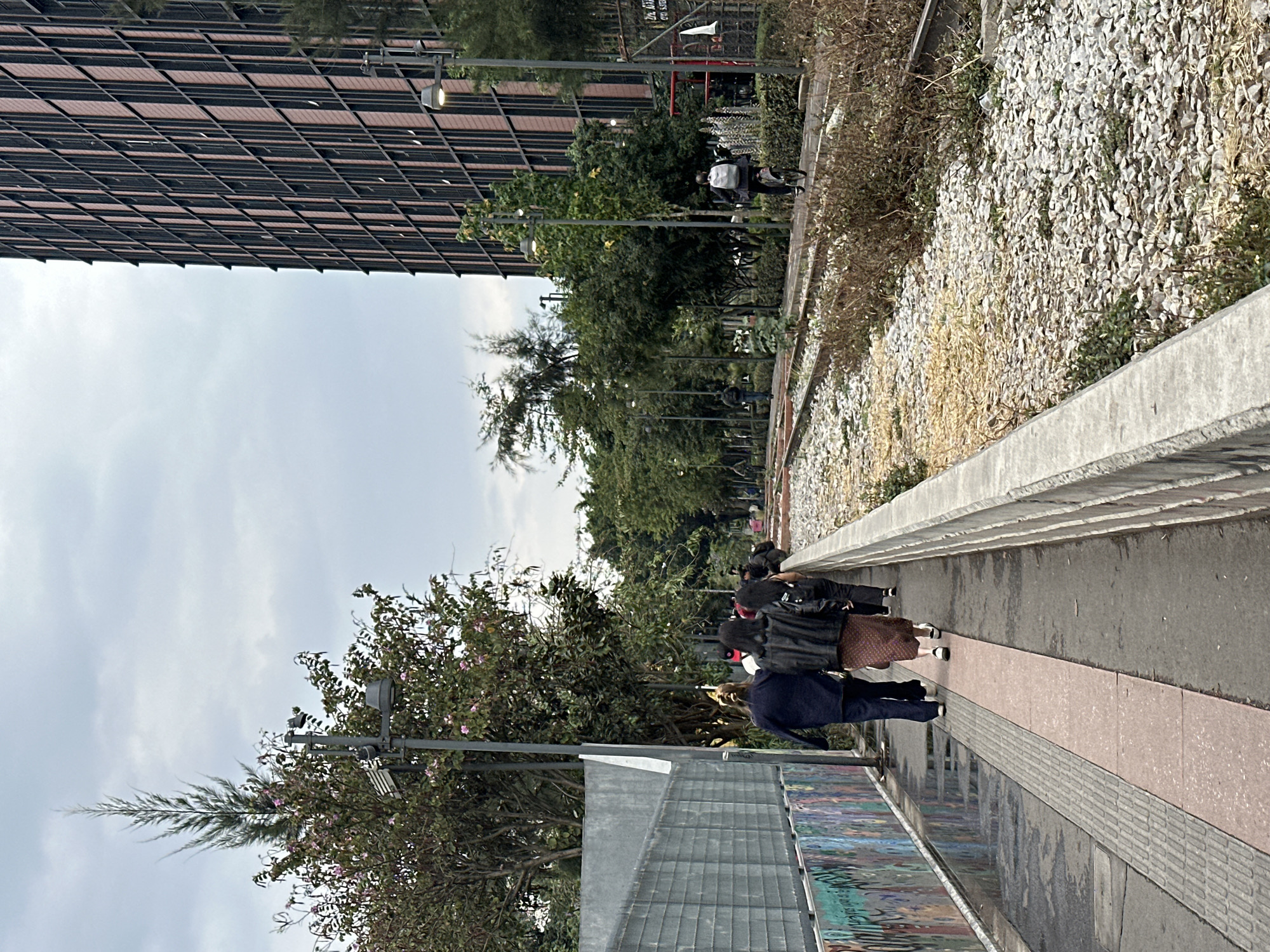 The image depicts a sidewalk in Mexico City, Mexico, adjacent to a building with a dark, grid-like facade. Several people are walking along the sidewalk, heading away from the viewer. To the right of the sidewalk, there is a raised area covered in light-colored gravel and vegetation. Streetlights line the sidewalk. The sky is overcast.