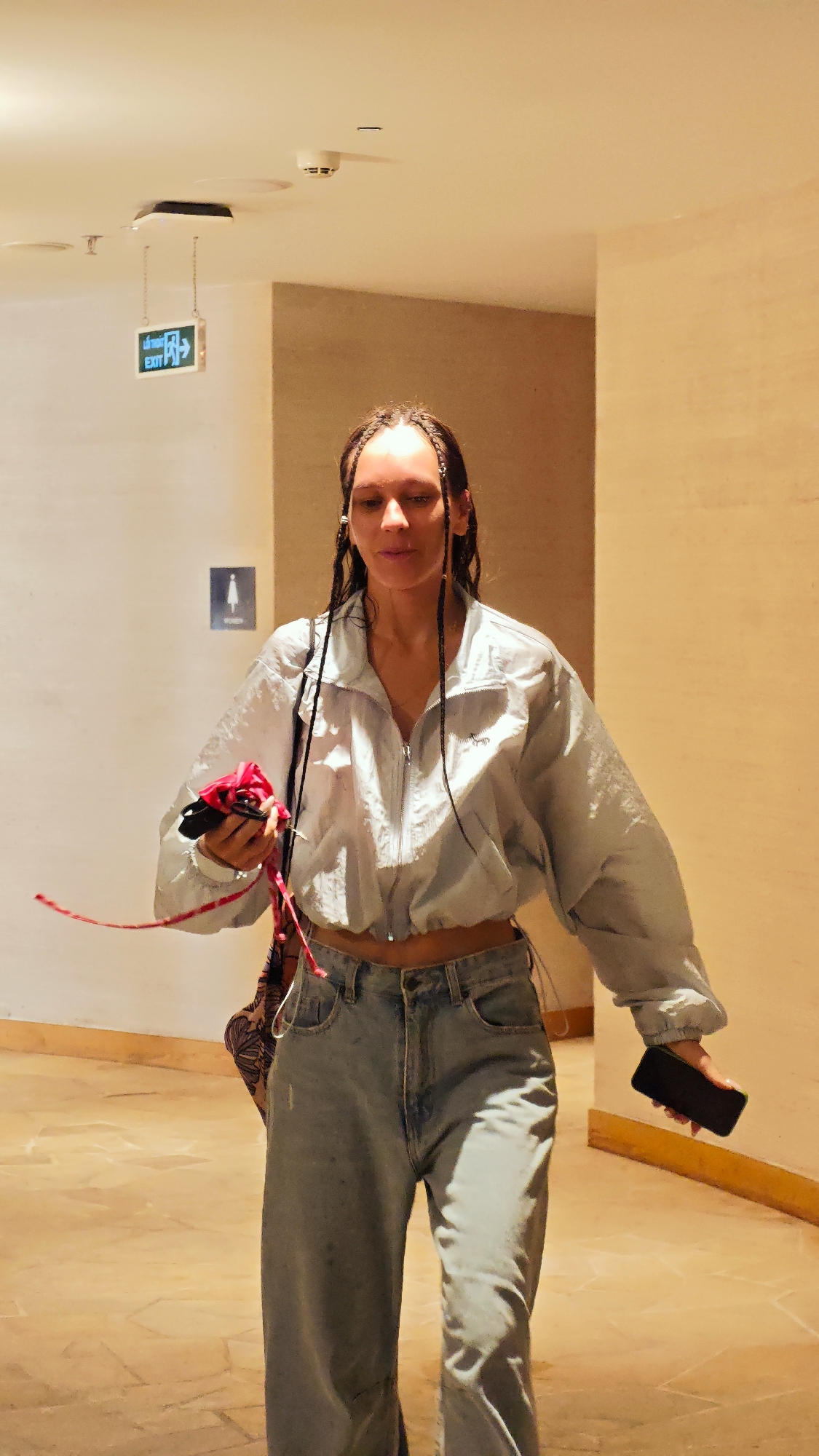 A woman with long, dark hair styled in numerous small braids is depicted walking through an indoor corridor. She wears a light blue or grey cropped windbreaker jacket with a zippered front, exposing a sliver of her midriff, paired with light-wash, wide-leg jeans. In her right hand, she carries a red ribbon or strap along with a piece of red and black fabric, possibly a face mask or small item. A black smartphone is held in her left hand.
The setting appears to be a well-lit hallway or lobby, characterized by warm, beige-colored walls and a matching tiled floor. The white ceiling features several fixtures, including a light, a smoke detector, and another electronic device. A green illuminated "EXIT" sign, displaying an arrow pointing right and text in both English and what appears to be another language, hangs from the ceiling. Further along the wall, a "WOMEN" restroom sign, indicated by a female stick figure, is visible. The artificial lighting gives the space a bright, inviting atmosphere. The woman's expression is neutral, and she appears to be moving purposefully.