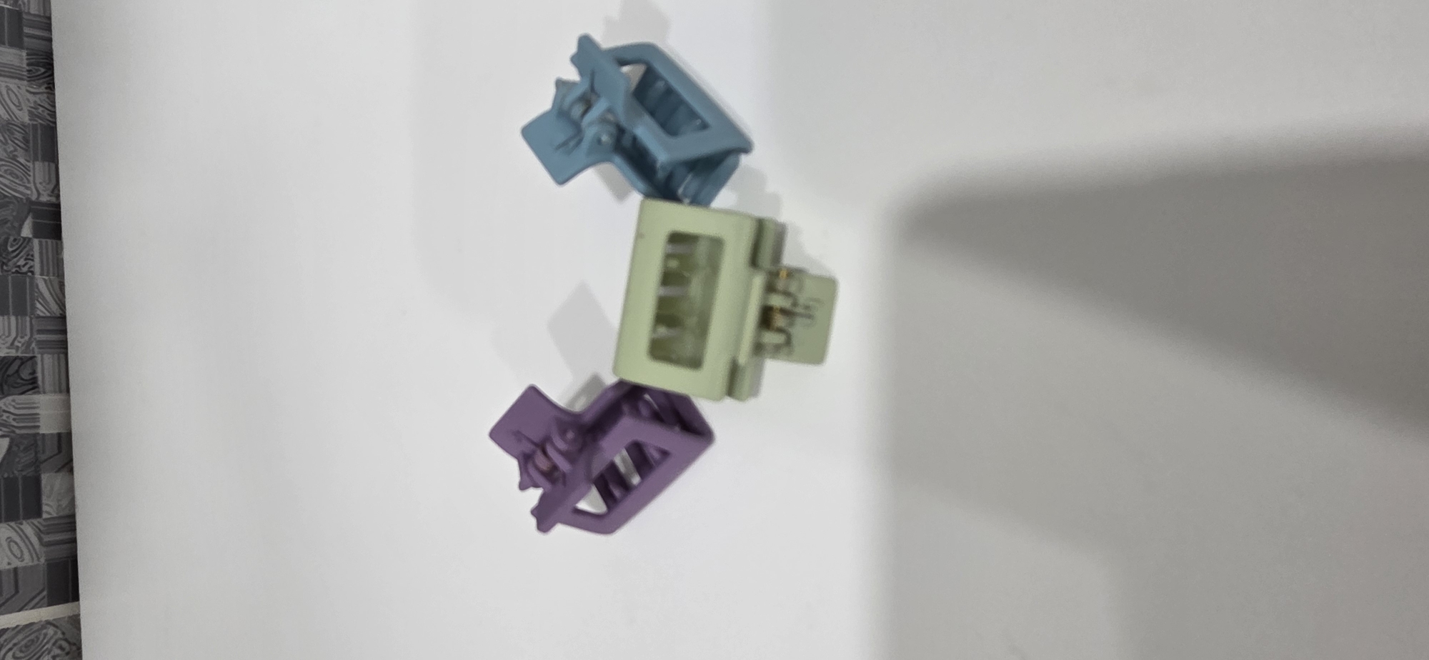 The image displays three small plastic hair claw clips on a flat, white surface. One clip, colored purple, is positioned on the left. A second clip, light green, is centrally located and slightly lower than the purple one. The third clip, light blue, is situated on the right. All three clips are oriented with their hinged mechanisms visible. Soft shadows are cast beneath each clip and extend faintly across the lower half of the white surface. The background, in the upper portion of the image, features a blurred, patterned surface, appearing to be composed of gray, black, and white tiles with abstract or geometric designs. The overall environment appears to be an indoor setting.