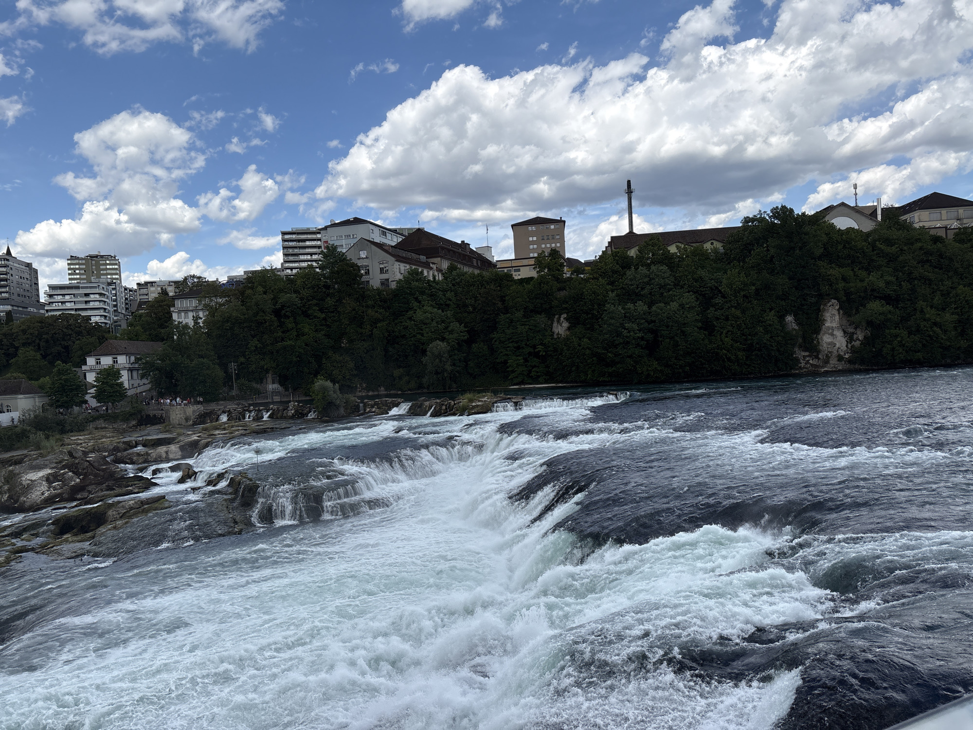 The image shows a wide, powerful waterfall cascading over dark, jagged rocks. The water is a frothy white and blue, with strong currents and turbulent waves. In the background, a lush green hillside is covered in dense trees. Above the trees, a line of buildings, some tall and modern, others older with tiled roofs, are visible against a bright blue sky dotted with fluffy white clouds. A tall, industrial-looking tower with an antenna stands out amongst the buildings on the right side of the frame. The scene appears to be a natural landscape integrated with an urban environment. This is likely the Rhine Falls, located near Dachsen, Switzerland, a significant natural landmark and tourist attraction.