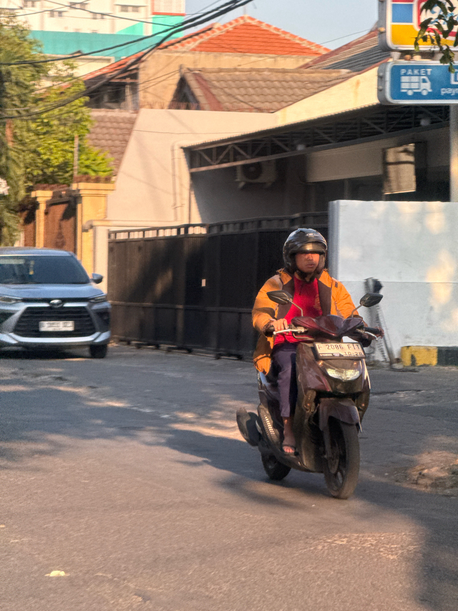 In Surabaya, Indonesia, a person wearing a dark helmet and an orange and yellow jacket rides a dark-colored motorcycle on a paved road. The rider is also wearing a red shirt and dark blue pants, with sandals visible on their feet. The motorcycle has a license plate reading "F 2086 FIT". In the background, a silver car is parked on the left side of the road. Behind the car and the motorcycle, there are houses with tiled roofs, a dark metal fence, and trees. A signpost with "PAKET" and a truck icon is visible on the right side of the image. The scene is illuminated by the warm light of either sunrise or sunset.