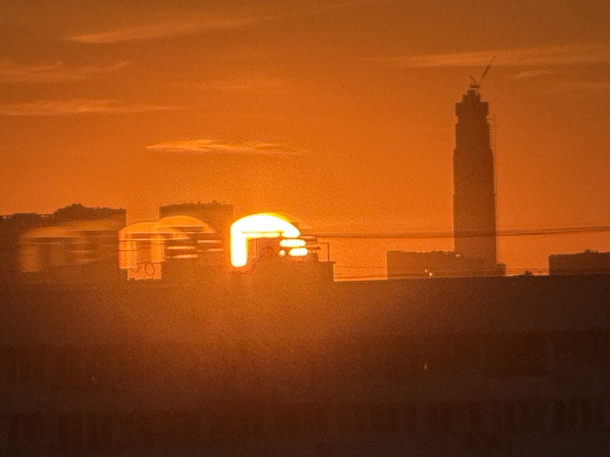 The original image is a medium shot of a city skyline at sunset. The sky is a vibrant orange, with a few wispy clouds. The sun, a bright white orb, is partially obscured by a building on the left side of the frame, casting a warm glow across the scene. In the center of the image, a tall skyscraper, possibly under construction, dominates the skyline. Its dark silhouette stands out against the fiery sky, and a crane is visible at its peak. To the left of the skyscraper, a row of lower buildings forms the cityscape. The foreground is dominated by a dark, silhouetted area, likely representing more buildings or a lower elevation of the city. The overall impression is one of a bustling urban environment bathed in the dramatic hues of a sunset.
