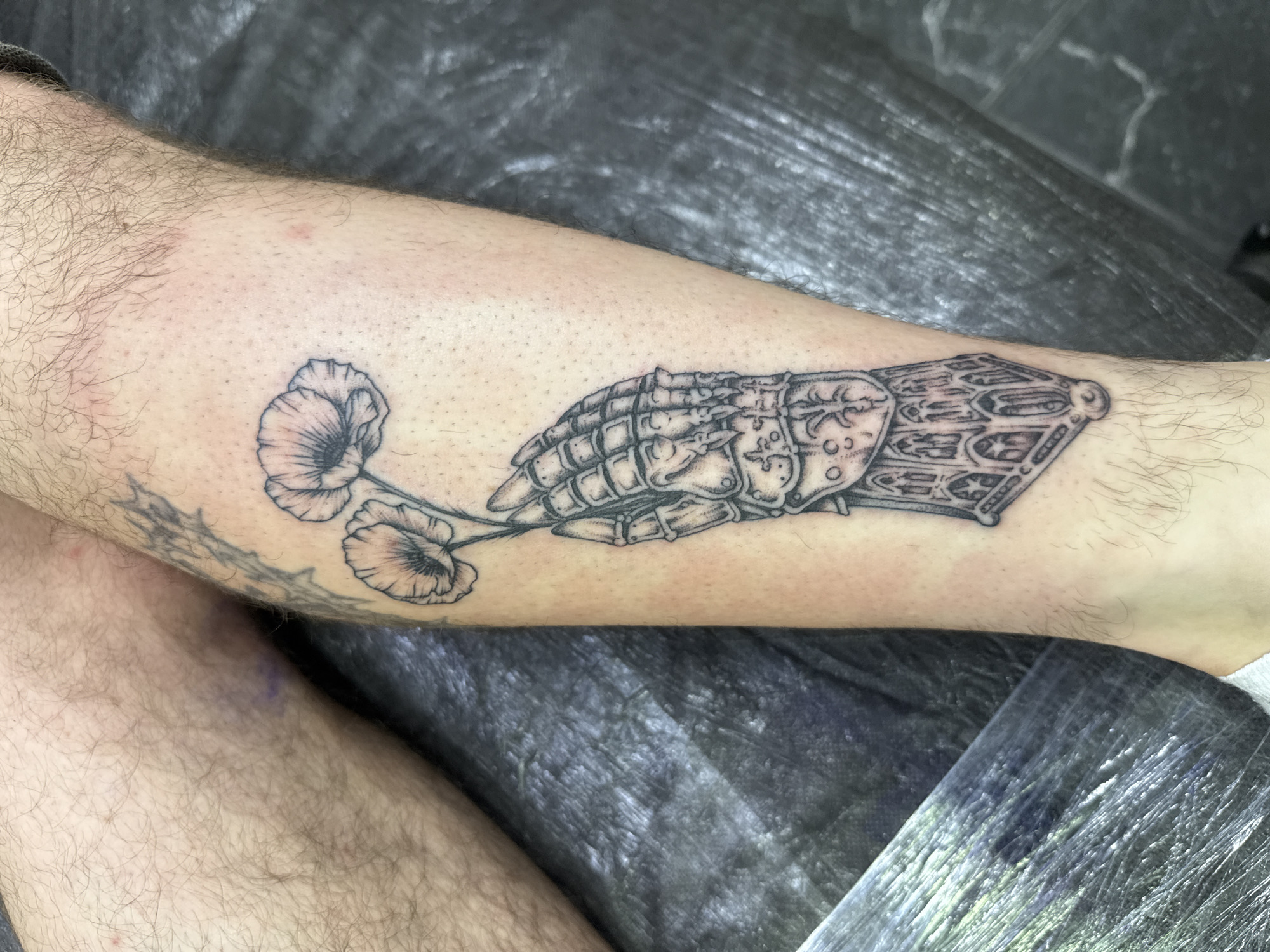 A black and white tattoo of a medieval gauntlet holding two poppies is depicted on the lower leg of a person. The tattoo is detailed, showing the intricate segments of the gauntlet and the delicate petals of the flowers. The surrounding skin is lightly tanned and has visible hair. The background is a dark, textured surface, possibly a tattoo parlor mat. There are no other people or discernible objects in the immediate vicinity of the tattoo. The location is unknown and cannot be determined from the image.