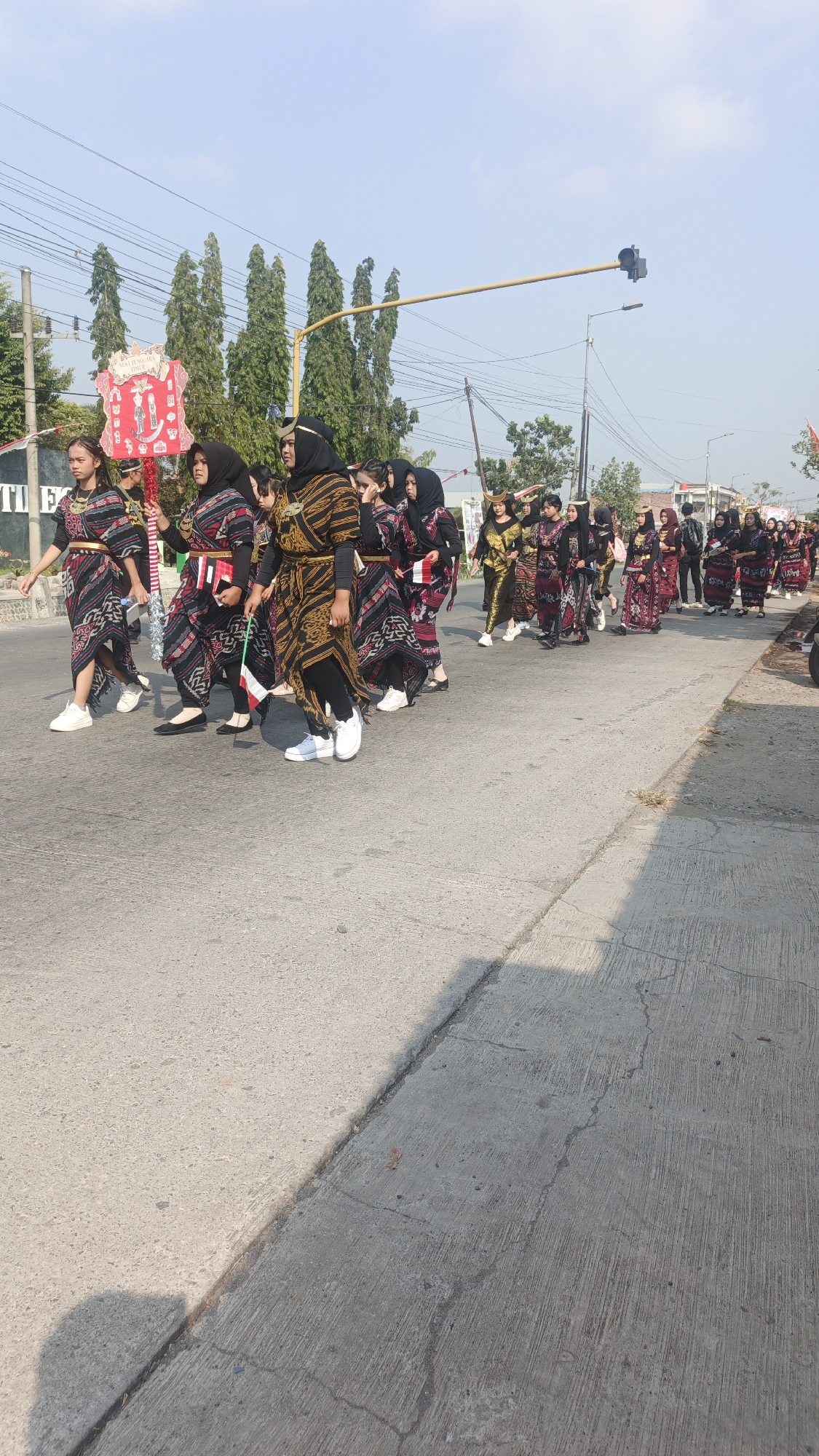 In Sumengko, Indonesia, a group of young women in traditional attire, featuring black headscarves and patterned dresses with gold accents, are participating in a parade. They are walking on a paved road under a clear blue sky, with tall trees lining the background. Some of the women are holding small Indonesian flags, while others are carrying a large, rectangular banner with red and white elements. The parade appears to be moving from left to right across the frame. Traffic lights and overhead wires are visible, suggesting an urban or semi-urban setting. The overall scene suggests a cultural or national celebration.