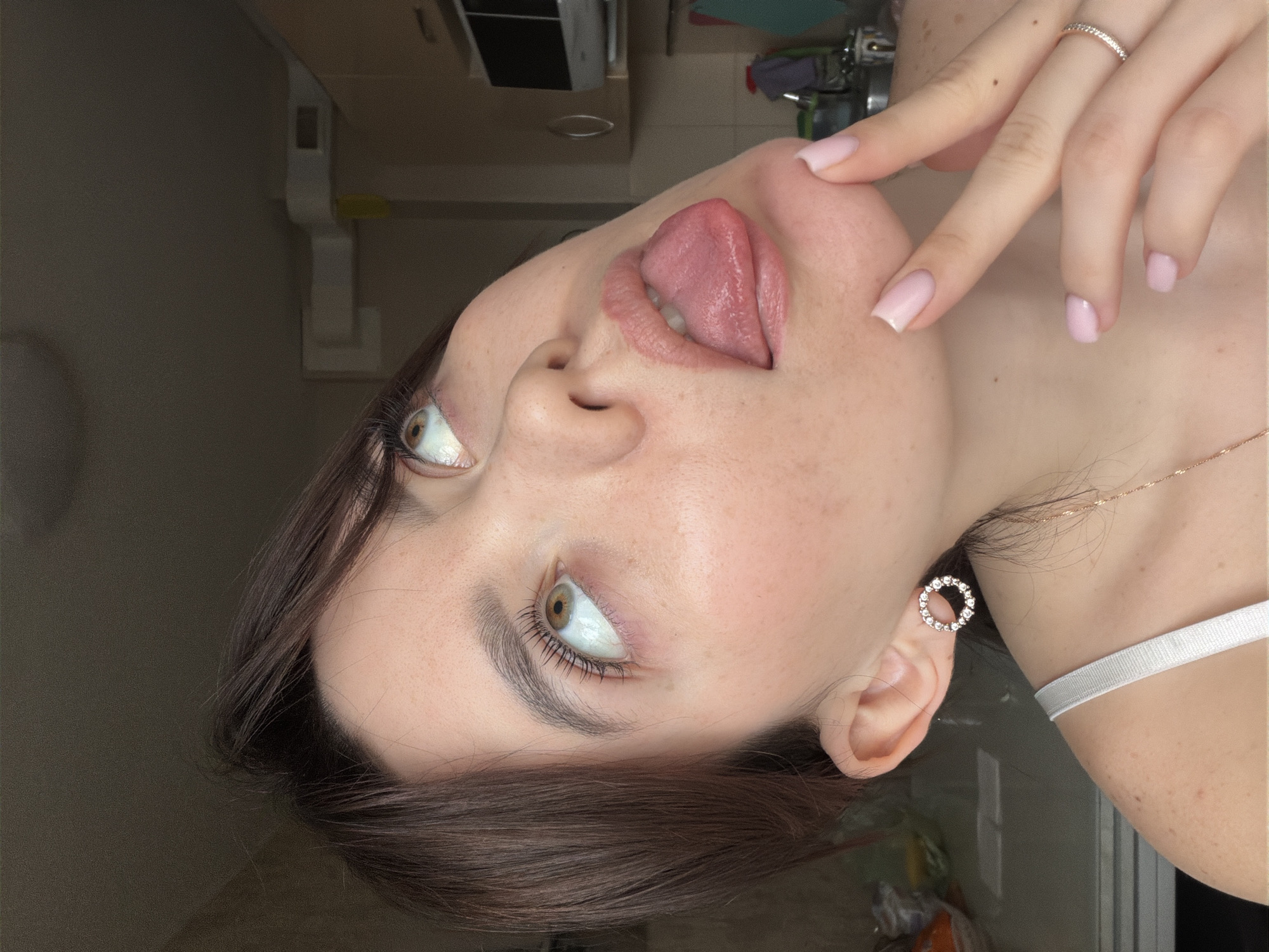 A close-up, angled photograph depicts a woman with light skin, brown hair, and light pink manicured nails, sticking her tongue out and touching her cheek with her fingers. She is wearing a ring on her finger, a necklace, and a circular earring. The background appears to be a kitchen or bathroom, featuring a sink, faucet, and cabinets. The setting could potentially be within an apartment or house in the city of Chaikovsky, Russia.