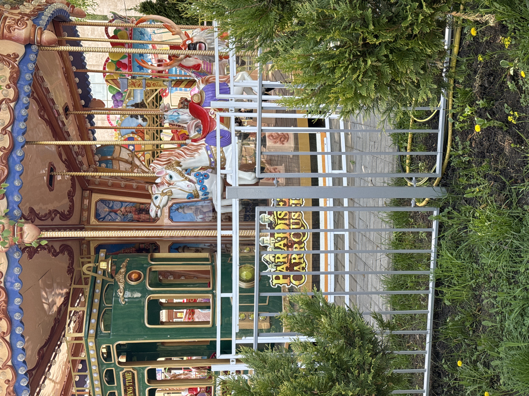 The image captures a carousel, identified by the sign "Paradise Carousel," behind a metal fence. The carousel features elaborately decorated horses and a green train car, indicative of a classic amusement park attraction. The surrounding environment includes greenery, suggesting the carousel is located in a park or recreational area. The image appears to be taken outdoors during the day, with natural lighting. The location is tagged as Unknown, Unknown, indicating an unspecified city and region.