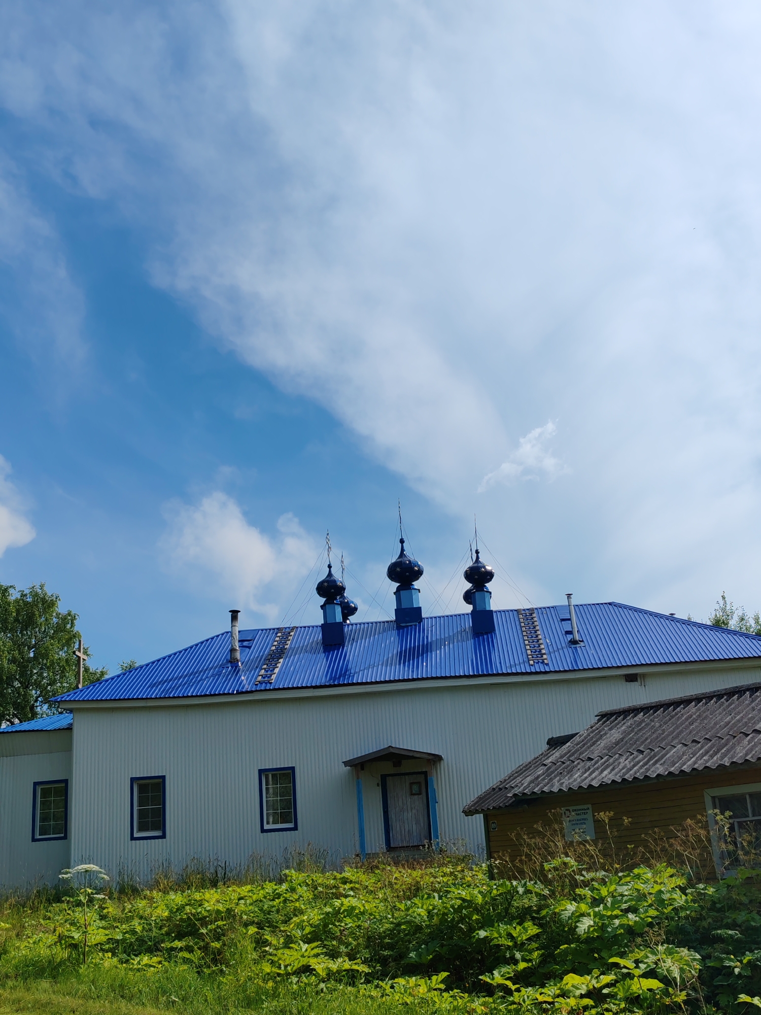 The image shows an exterior view of a white church with a bright blue, corrugated metal roof, adorned with several dark blue onion domes topped with crosses. The church has multiple windows with white frames and blue trim. In front of the church, there is a smaller, older-looking wooden building with a gray, corrugated roof. The area is surrounded by lush green vegetation, including tall grasses and leafy plants, suggesting a rural or semi-rural setting. The sky is partly cloudy with a bright blue expanse. The scene appears to be located in Chuchloma, Russia, indicated by the context provided. There are no people visible in the image.