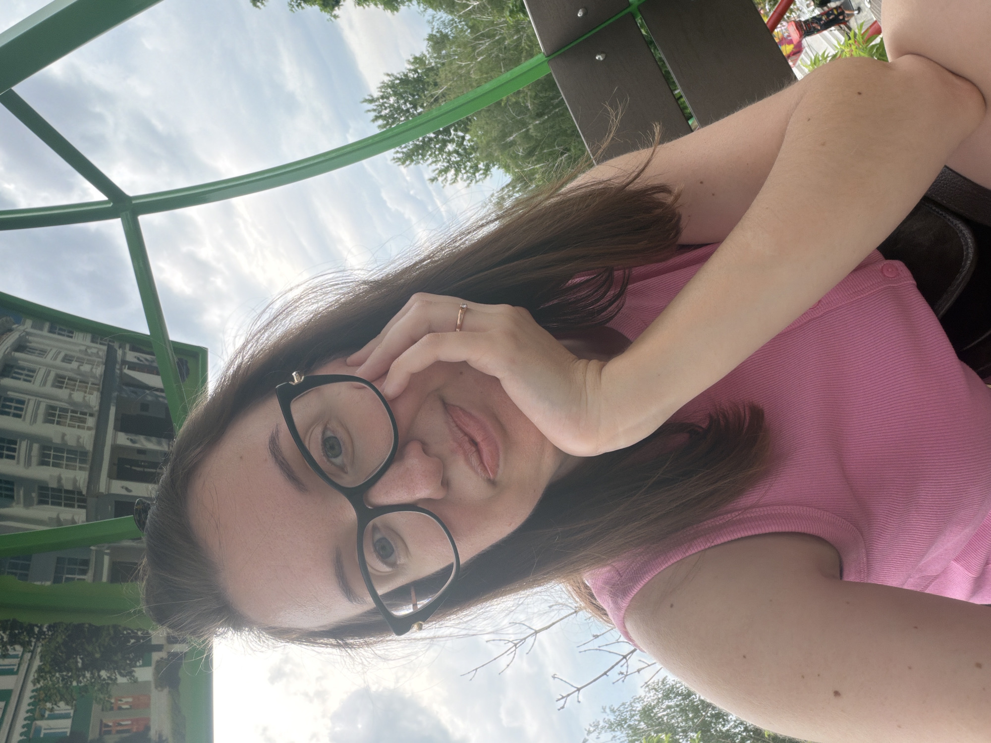The image is a medium close-up selfie of an adult female, positioned slightly off-center to the left, looking directly at the camera with a subtle, closed-mouth smile. She has long, dark brown hair parted slightly to the left, and is wearing black-framed glasses with gold accents near the temples. Her skin tone is light, and faint freckles are visible across her nose and cheeks. Her lips are a light natural pink. She wears a pink ribbed sleeveless top. Her right hand, adorned with a simple gold ring featuring a single small clear stone on the ring finger, supports her chin. Her left arm is partially visible.
The background is dominated by a bright green angular metal structure, likely part of a playground or park seating, which frames the upper and right portions of the scene. In the upper left, within the green frame, a reflective surface or sign displays an image of multi-story buildings featuring classical architecture with columns, arches, and light-colored facades, interspersed with some greenery. The sky above is overcast with white-grey clouds. Lush green deciduous trees are visible in the mid-background, particularly to the right of the subject, and some bare tree branches appear on the left. A brown wooden park bench with visible metal studs is partially obscured behind the subject's right arm.
Based on the architectural style depicted in the background image and the overall park setting, the location is identified as the Alexandria Dendrological Park in Bila Tserkva, Kyiv Oblast, Ukraine.