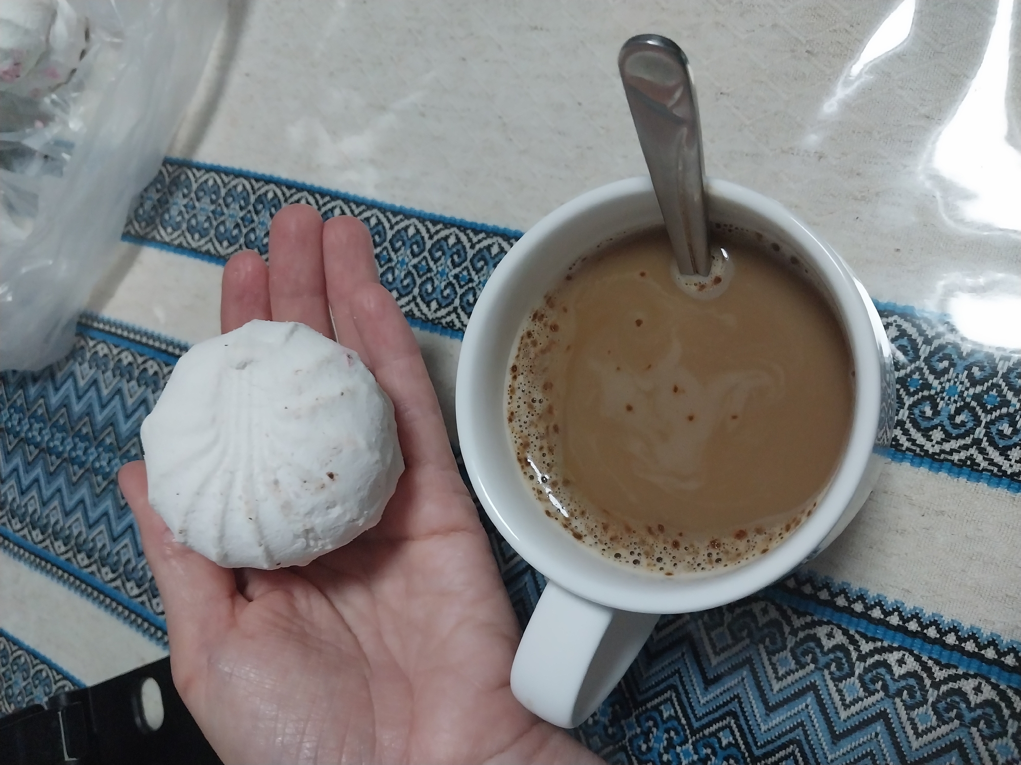 A close-up, overhead shot shows a person's hand holding a white marshmallow over a white mug of coffee. The marshmallow has a textured surface with some brown speckles. The coffee in the mug is light brown with a foamy head and a silver spoon resting in it. The background consists of a patterned tablecloth with blue and white geometric designs. The scene is domestic, possibly a breakfast or snack setting. The image quality is slightly grainy, suggesting it was taken in low light conditions. The location is not specified within the image content.
