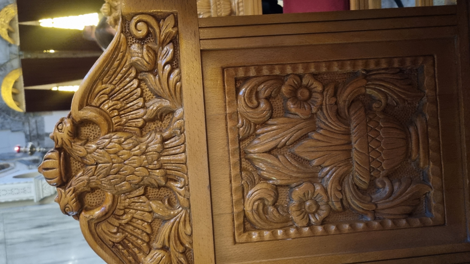 A close-up, eye-level, medium shot captures the intricate wooden carvings of what appears to be a throne or a significant piece of furniture within a religious building. Dominating the upper portion of the frame is a detailed relief of a double-headed eagle, an emblem often associated with Byzantine history and the Greek Orthodox Church. The eagle is crowned, with its wings spread and feathers meticulously carved. Below this, a rectangular panel features a carved floral motif, centered around an ornate vase overflowing with leaves and flanked by two stylized flowers. The wood is a warm, medium brown, with visible grain and subtle signs of wear. The background is blurred, showing architectural elements of what could be a church in Thessaloniki, Greece, with marble columns and arched openings, as well as glimpses of yellow light filtering through, suggesting stained glass or illuminated windows. A sliver of red fabric is visible on the right, possibly part of another seat or altar cloth. The overall impression is one of rich, historical craftsmanship within a sacred space.