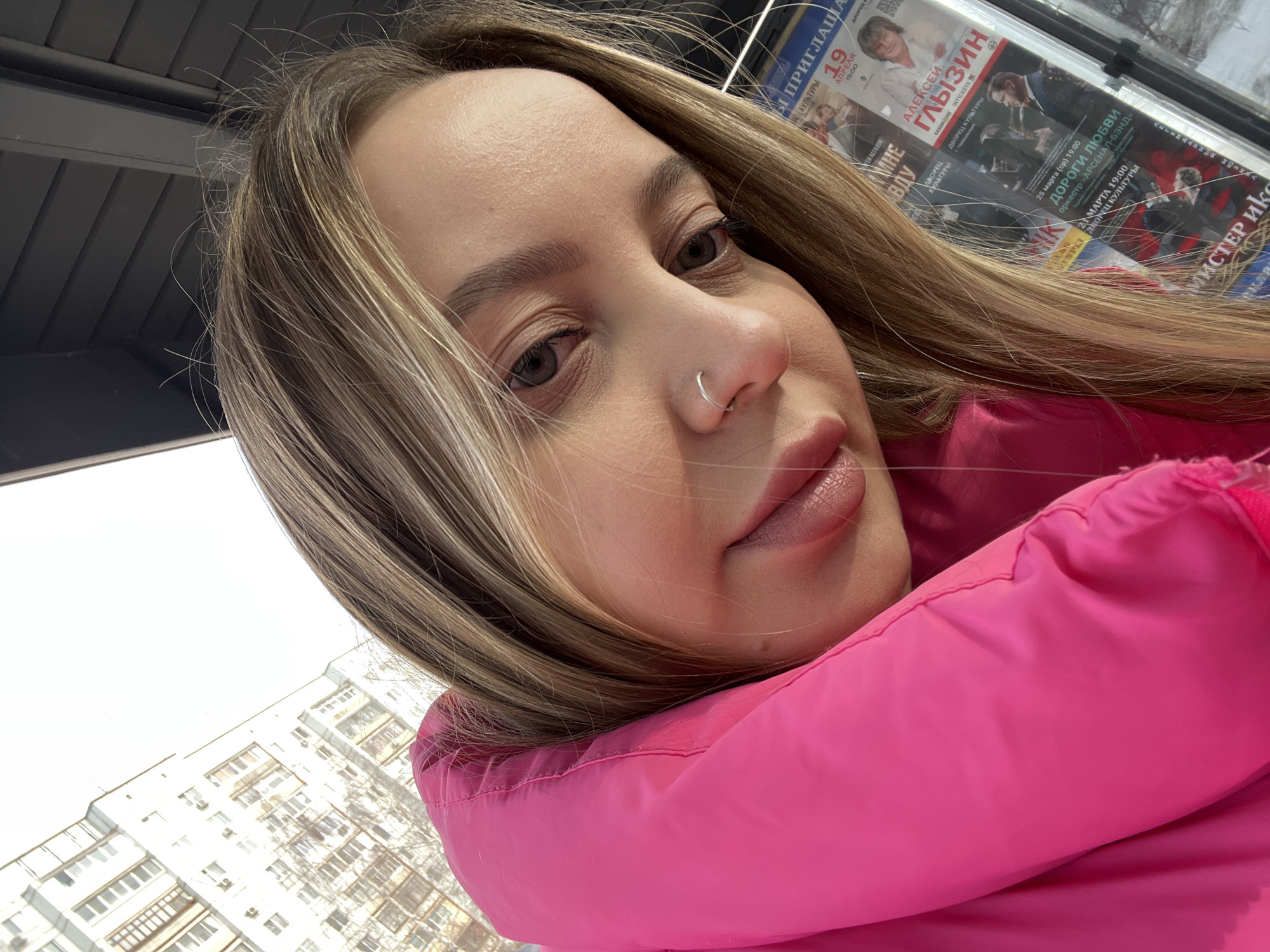 A close-up, slightly angled selfie depicts a young adult female with blonde hair, fair complexion, and light eyes, wearing a vibrant pink puffer-style jacket. She has a silver hoop nose ring in her left nostril and a small mole below her right eye. Her gaze is directed towards the camera.
In the background to the upper left, a large, multi-story residential building with numerous windows is visible, painted in a light color, under a bright, possibly overcast sky. To the subject's right, a glass-enclosed public display features several Russian concert posters. Prominently displayed is an advertisement for "АЛЕКСЕЙ ГЛЫЗИН" (Alexey Glyzin) scheduled for "19 АПРЕЛЯ 19:00" (April 19th, 19:00) at "ДВОРЕЦ КУЛЬТУРЫ" (Palace of Culture). Other posters announce performances on "25 МАРТА" (March 25th) by "оркестр «АРСЕНАЛ-БЭНД»" (Arsenal-Band orchestra) and on "21 МАРТА" (March 21st), both also at the "ДВОРЕЦ КУЛЬТУРЫ". Based on these specific concert advertisements for the Palace of Culture, the scene is located in the city of Chaikovsky, Russia, likely in the vicinity of the local cultural center.