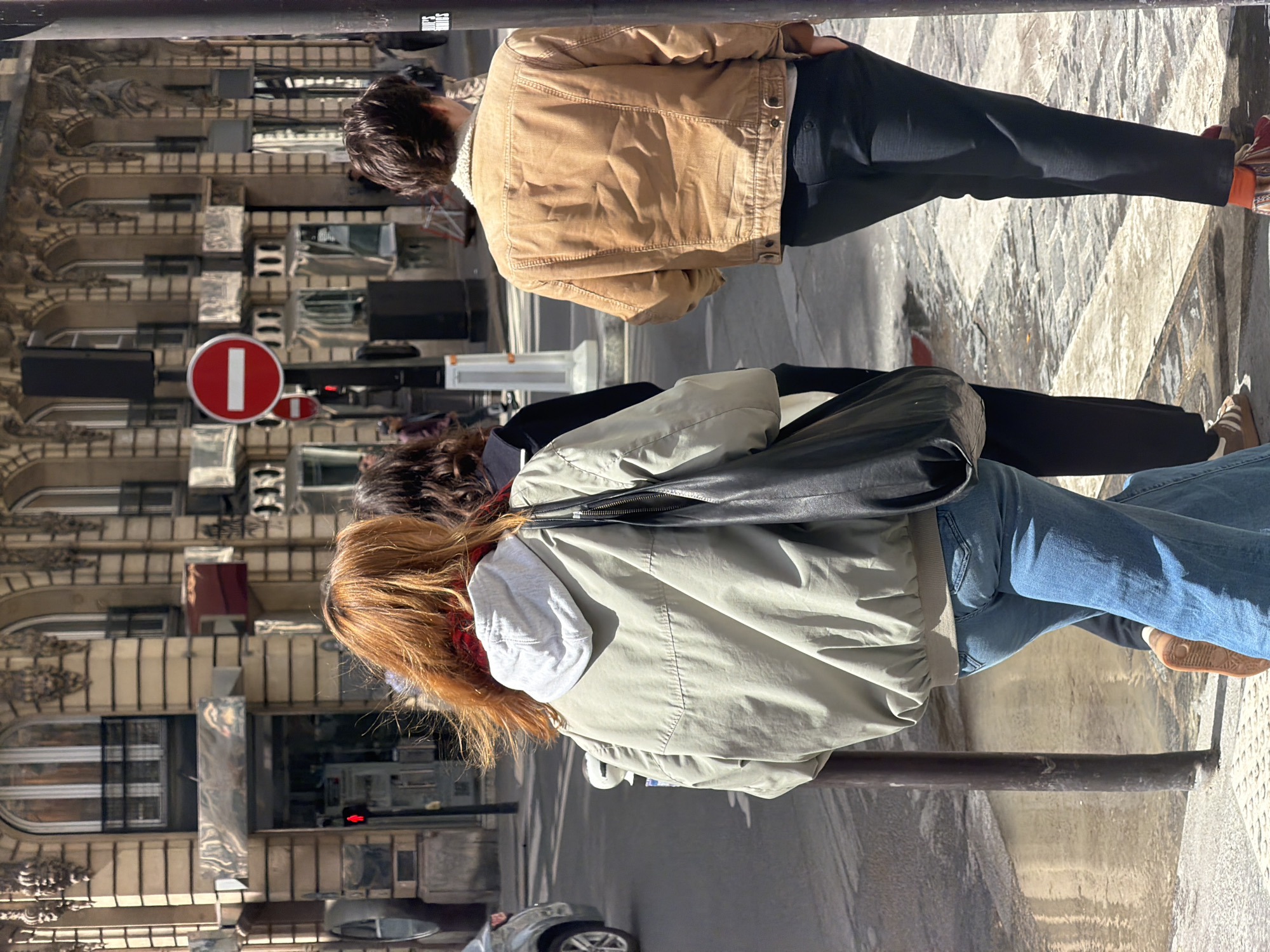 The image captures an urban street scene in Paris, France, on a bright, sunny day. Three individuals are seen from behind, walking along a patterned sidewalk.
The furthest person wears a tan or light brown jacket with a textured collar, possibly sherpa, and dark trousers. Their hair is dark brown.
The central figure is dressed in a light olive-green jacket, layered over a grey hooded sweatshirt, and blue jeans. They carry a black shoulder bag over their right shoulder and have reddish-brown hair.
The person closest to the camera is partially visible on the far left, showing only the back of their dark brown hair and a glimpse of a dark garment.
In the background, multi-story buildings with classic Parisian architectural details, including decorative facades and arched windows, line the street. A red circular traffic sign with a white horizontal bar, commonly indicating "No Entry" or "Do Not Enter," is prominently displayed on the left side. The strong sunlight creates clear shadows, suggesting a bright and clear day. A portion of a parked car is visible in the lower left corner. The individuals appear to be casually walking, but no specific activity or emotion can be discerned as their faces are not visible.
