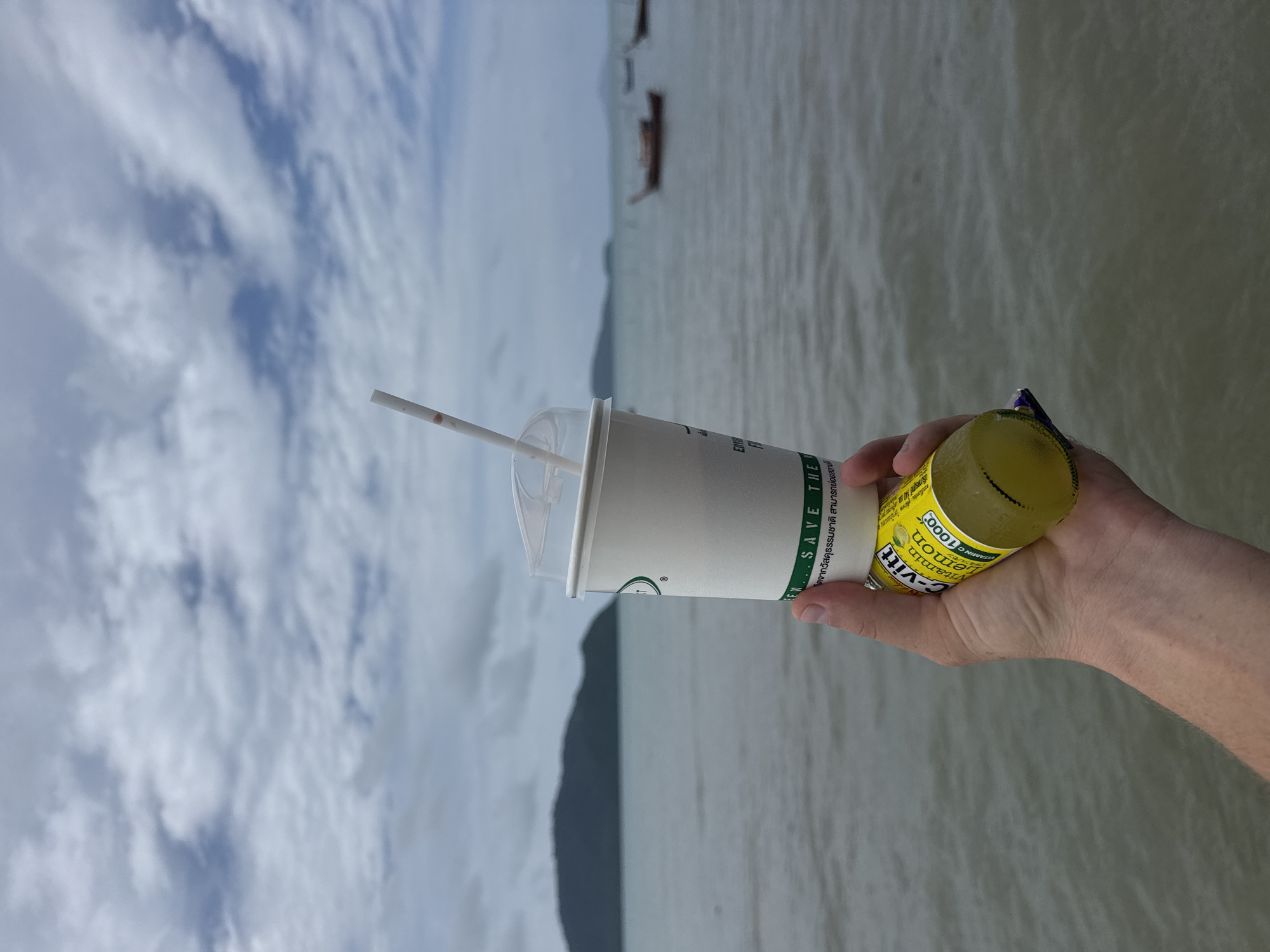 A hand holds a paper cup with a straw and a small bottle of "C-vitt Vitamin Lemon 1000" against a backdrop of the sea and a cloudy sky. The cup is white with green and black lettering that includes "SAVE THE E". The bottle is yellow and has text in what appears to be Thai script. In the distance, a green, hilly island is visible, along with several small boats on the water. The image was taken from the shore, possibly from a pier or beach in Rawai, Thailand. The water appears calm and slightly choppy. The lighting suggests an overcast day.