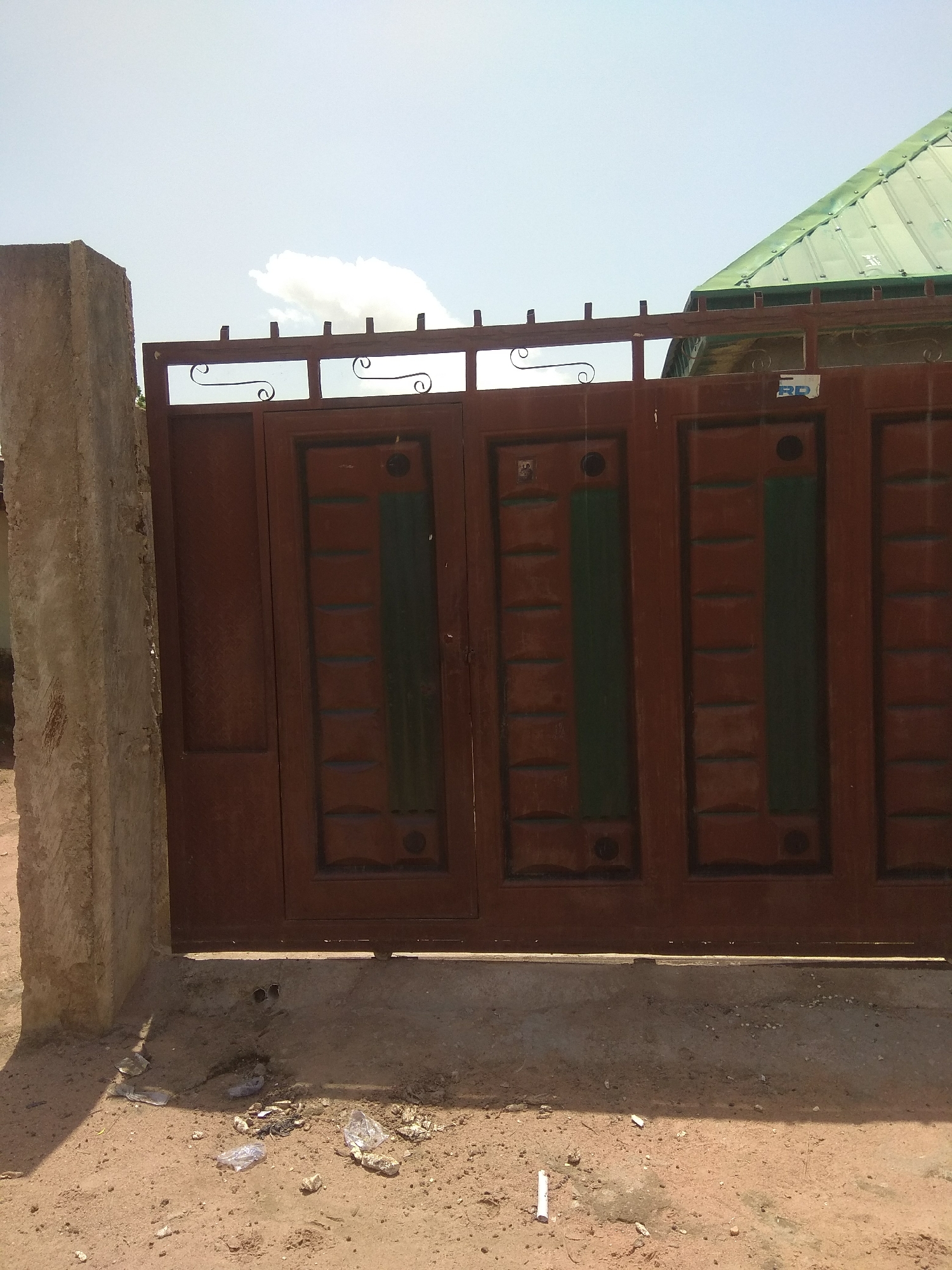 The image shows a close-up view of a brown metal gate in Jalingo, Nigeria. The gate is divided into three panels, each with a decorative rectangular inset featuring horizontal lines and a vertical green stripe. The top of the gate has a decorative metal railing with scrollwork. To the left of the gate, a concrete pillar stands prominently. The ground in front of the gate is sandy and littered with some debris and plastic bags. In the background, a green corrugated metal roof is visible, suggesting the presence of a building. The sky is bright and appears to be a clear day with a few scattered white clouds, indicating it is likely daytime. There are no people visible in the image. A small blue and white sticker with the letters "RP" is affixed to the upper right section of the gate.