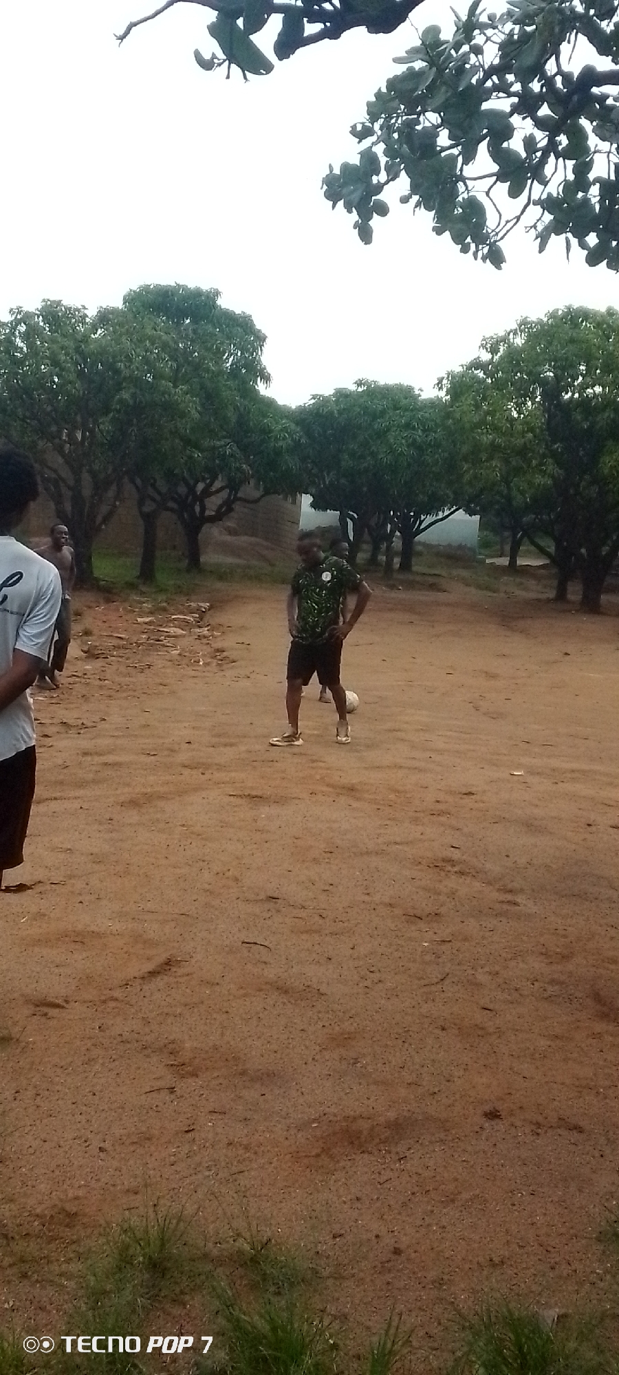 The image shows a group of people playing soccer in an outdoor dirt field. In the foreground, on the left, a person wearing a light-colored t-shirt and dark pants is partially visible, facing away from the camera. In the middle ground, a person in a green and black patterned t-shirt and black shorts is actively engaged with a soccer ball, which is visible in front of them. To the left of this player, another person is standing, shirtless, looking towards the soccer player. In the background, there are several trees with dense green foliage and a building structure that appears to be made of stone or brick. The ground is composed of dry dirt with some scattered debris and patches of green grass along the bottom edge. The sky is overcast, suggesting a cloudy day. The overall scene depicts a casual game of soccer in a rural or semi-rural setting.
