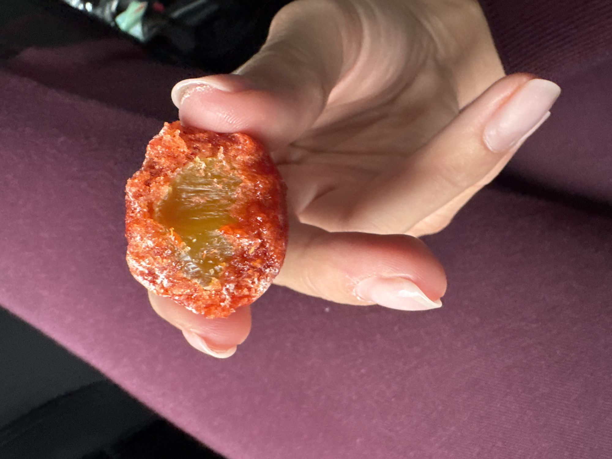 A close-up shot shows a person holding a spicy tamarind candy, possibly a "pelon pelo rico" variant, between their thumb and index finger. The candy is cylindrical with a hollow center filled with a yellow substance. The outer layer is coated in a bright red, granular seasoning. The person has light skin and manicured nails. The background features a dark burgundy fabric. The location is indeterminable from the image.