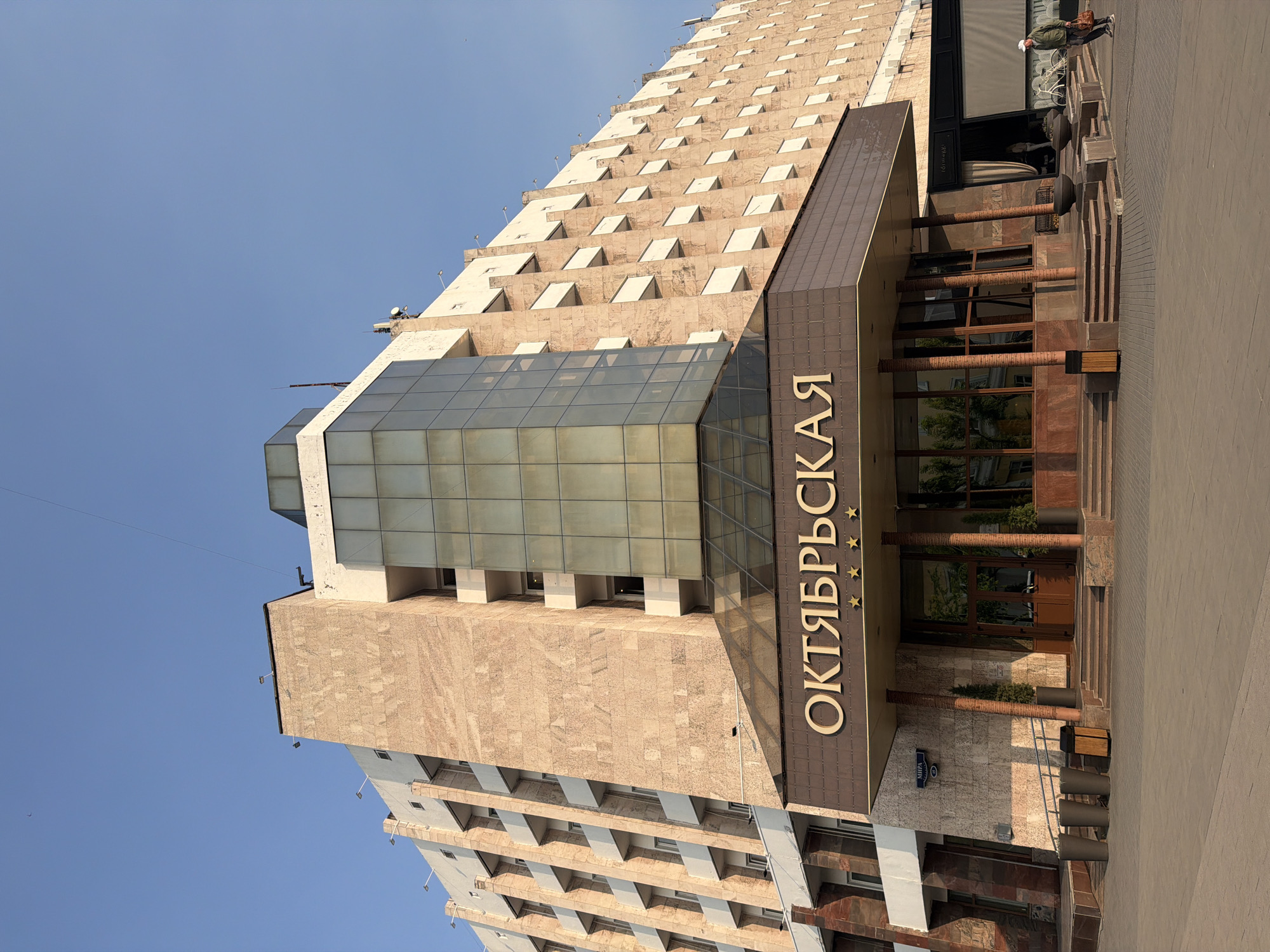 A wide shot captures the exterior of the Oktyabrskaya Hotel in Krasnoyarsk, Russia, under a clear blue sky. The imposing building features a light brown, textured stone facade with repeating rows of balconies. A modern glass-enclosed section rises from the center of the hotel, contrasting with the older architecture. The entrance is marked by a brown awning with the hotel's name, "ОКТЯБРЬСКАЯ," written in large gold letters, flanked by three gold stars on either side. Brown pillars support the awning, and stairs lead up to the main entrance. To the right of the entrance, a person wearing a white cap, green jacket, and dark pants walks away from the camera, carrying a brown bag. Bicycles are parked to the right of the hotel entrance, and planters with small trees are placed near the steps. The ground in front of the hotel is paved with light-colored tiles.