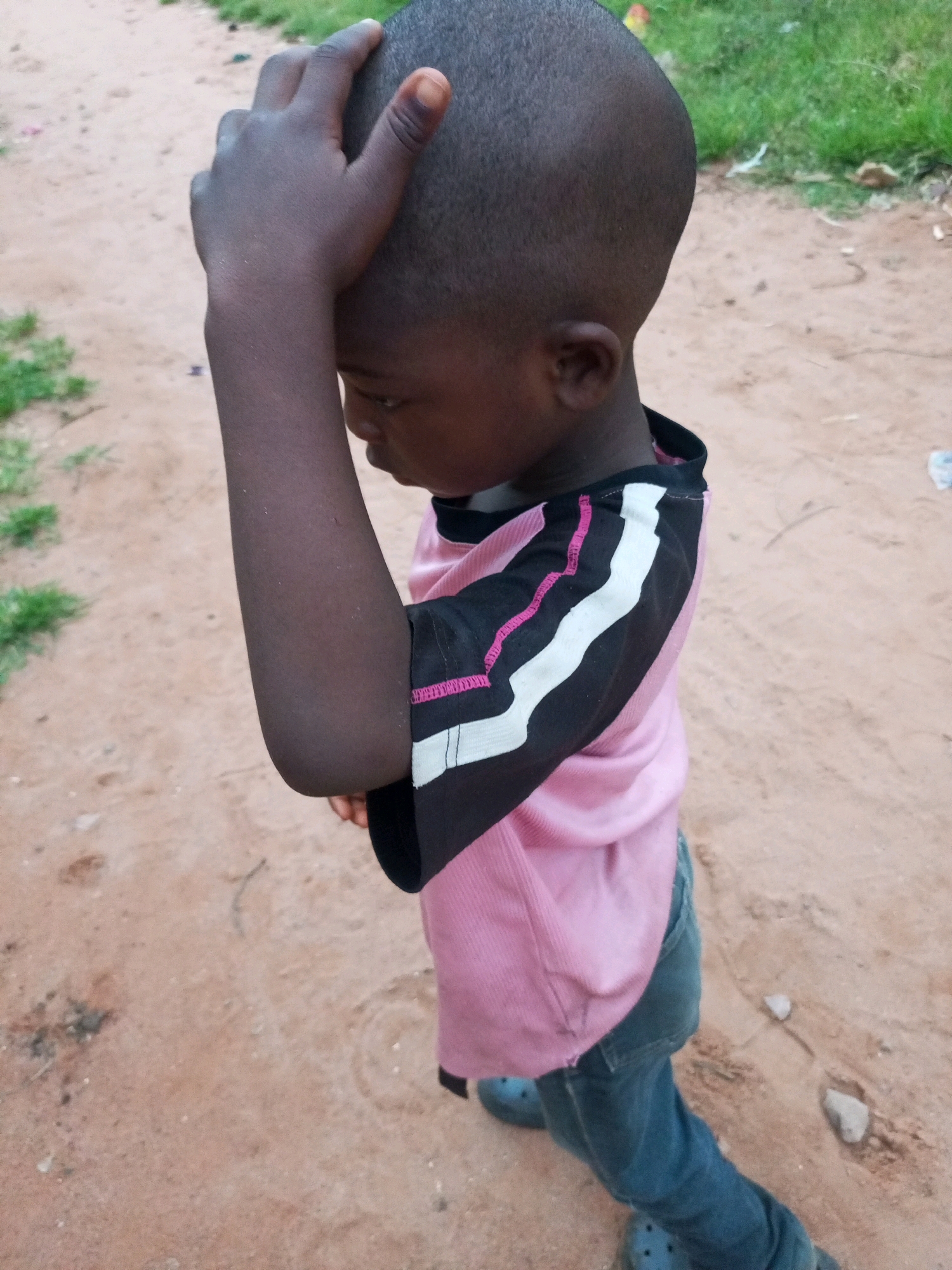 A young Black child, facing right and looking down, stands on a dirt path in Tempure, Nigeria. The child has a shaved head and is wearing a pink t-shirt with black and white stripes on the sleeves, and blue jeans. Their right hand is touching their head. The environment consists of a dirt path with patches of dry grass and some scattered debris. The lighting suggests it is daytime.
