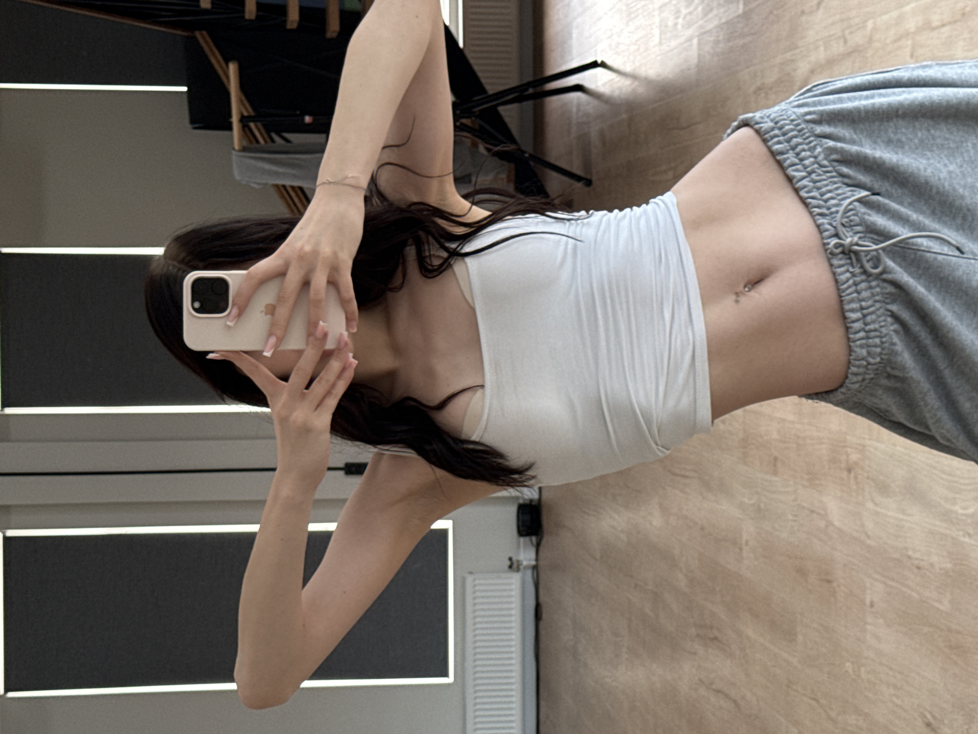 The image shows a young woman taking a mirror selfie in what appears to be a dance studio or gym. She is wearing a white strapless crop top and grey sweatpants. Her hair is dark and long, falling around her shoulders. Her left arm is raised, holding a light-colored smartphone to take the photo. Her right arm is bent, with her hand near her face. The background shows a wooden floor, a radiator, and some vertical blinds or paneling with light strips. In the reflection of the mirror behind her, there's a glimpse of some equipment, possibly dance barres or racks. The lighting suggests it's daytime. No text is visible in the image.