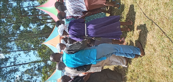 This image captures a group of people gathered outdoors in Burundu, Kenya, under bright, sunny conditions, likely midday or early afternoon given the prominent shadows on the grass. The scene features several adults, both men and women, attending what appears to be a community or social event.
In the background, two colorful tarpaulin structures, serving as shade covers or temporary shelters, are visible. One is pink and green, and another is light blue and orange. Tall trees with green foliage line the horizon, indicating an open, natural setting.
The people are positioned in what seems to be rows, mostly facing away from the camera or in profile. Notable attire includes a woman in the midground wearing a long, deep purple dress with a white head covering, and a measuring tape visible around her waist. In front of her, a man wears a light blue shirt over light-colored trousers. Other individuals are dressed in various casual and traditional clothing.
The ground is covered in dry-looking green grass, and strong shadows cast by the people indicate intense sunlight. A long, dark, thin object, possibly a cable or rope, lies on the grass in the lower right portion of the frame. The overall atmosphere suggests a communal outdoor gathering. There is no clearly readable text visible within the image beyond the unreadable markings on the measuring tape.