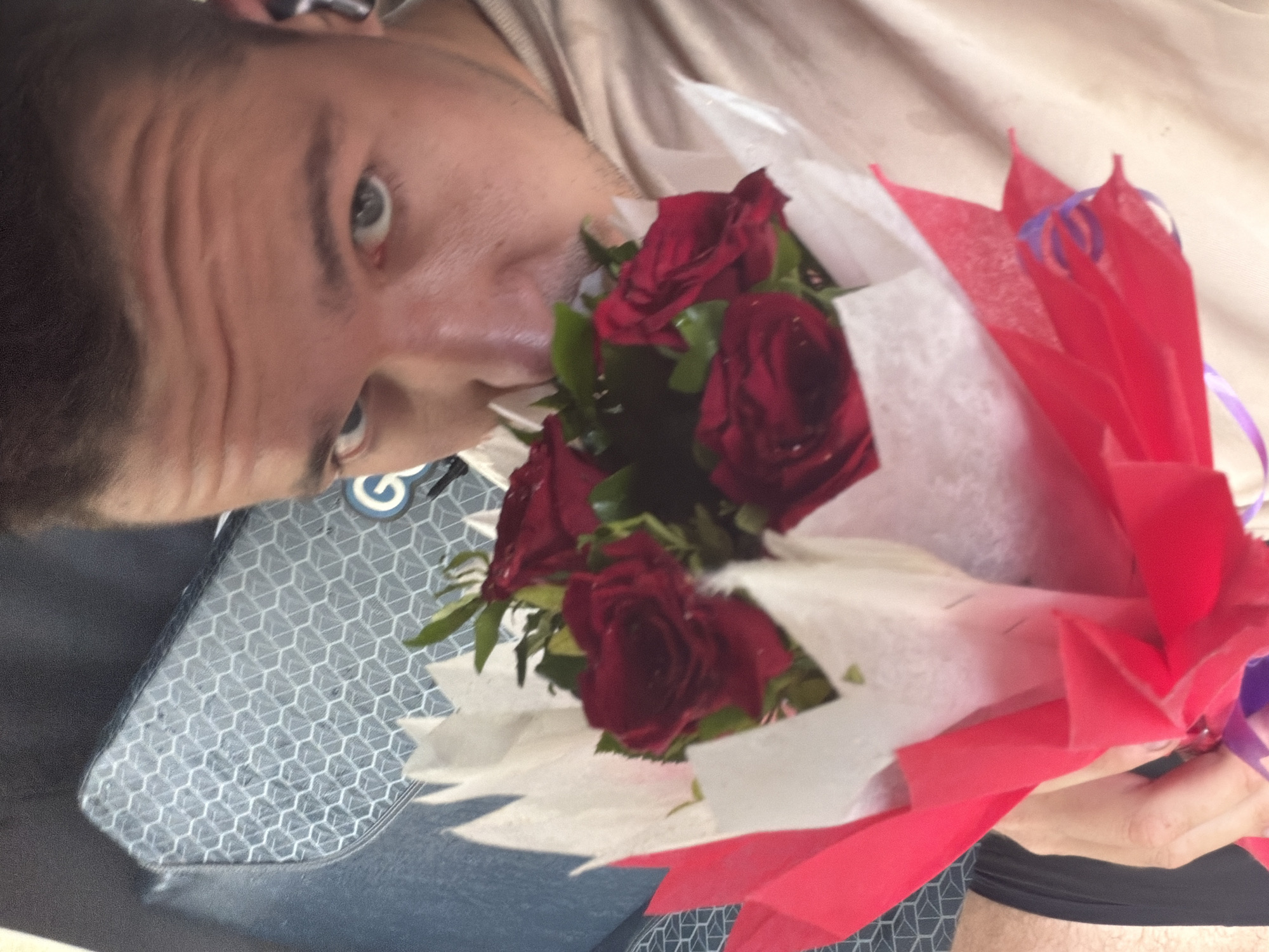 The image shows a close-up of a person holding a bouquet of red roses, with their face partially obscured as they smell the flowers. The person appears to be male, with short dark hair and blue eyes visible. They are wearing a light-colored shirt and have an earbud in their right ear. The bouquet is wrapped in red and white paper, with a purple ribbon tied around it. In the background, a textured surface with a hexagonal pattern is visible, possibly part of a seat. The lighting suggests an indoor or shaded outdoor setting. There is no indication of the city of Kanpur, India, in the image.