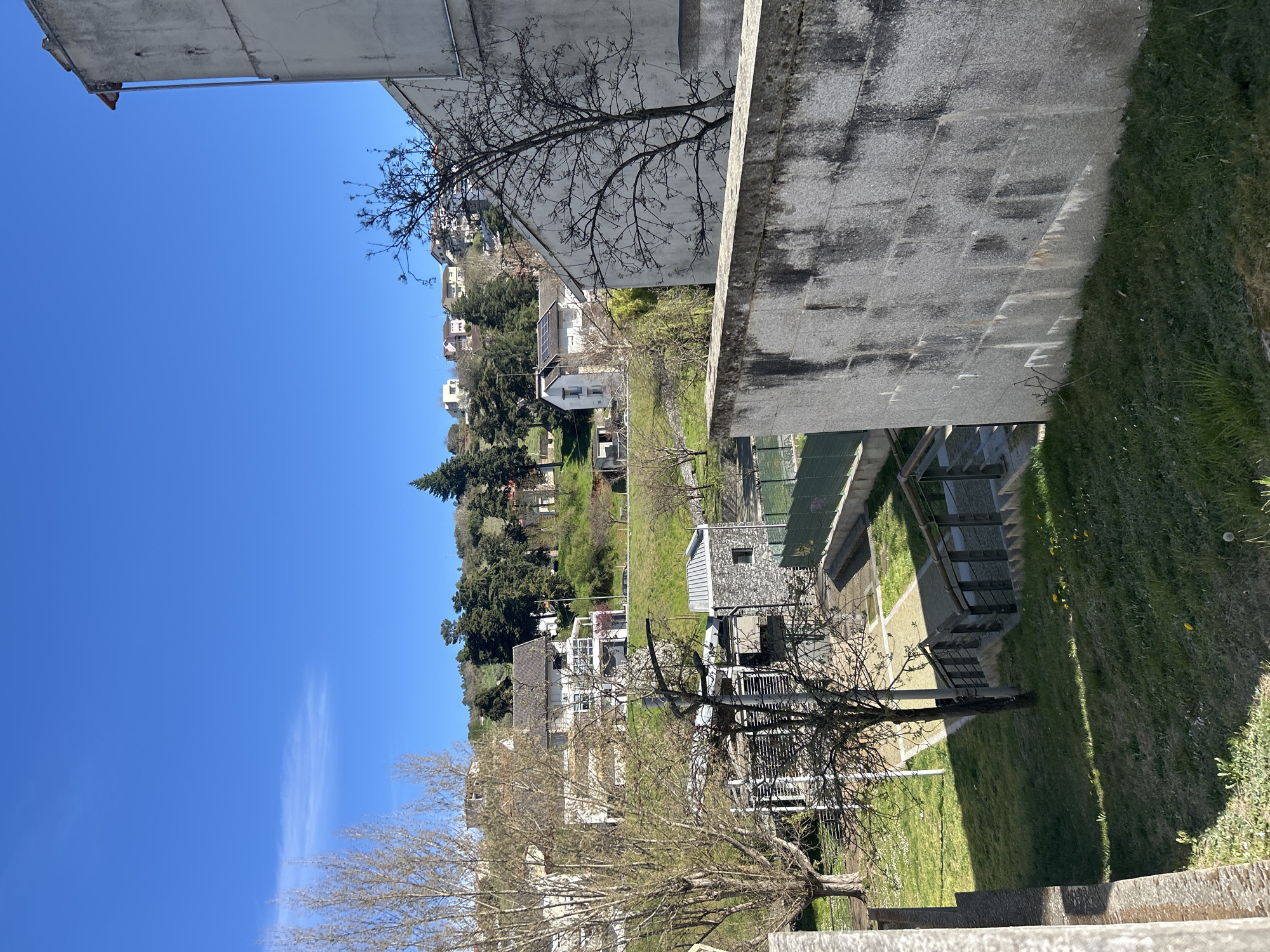 This image presents an elevated, static outdoor scene in Bragança, Portugal, capturing a residential and green landscape under a bright, clear blue sky with a single faint wispy cloud in the upper left. The immediate foreground on the right features a large, light grey concrete structure or wall, exhibiting prominent dark vertical staining from weathering, which casts a defined shadow onto a grassy slope. A bare-branched deciduous tree is positioned in front of and to the right of this wall. Below and to the left, a set of concrete stairs with dark metal railings descends into a lower-lying area of grass and paved pathways. Adjacent to the stairs and further left, a modern building with a grey stone facade and a metal roof is visible, situated next to a green fence that likely encloses a recreational court. The background reveals a hilly terrain ascending into the distance, characterized by a mix of bare and evergreen trees and multiple scattered residential houses. These houses are predominantly white or light-colored with visible tiled roofs. One specific house on the left side of the background features solar panels installed on its roof. No individuals, explicit actions, or interactions are discernible within the frame.