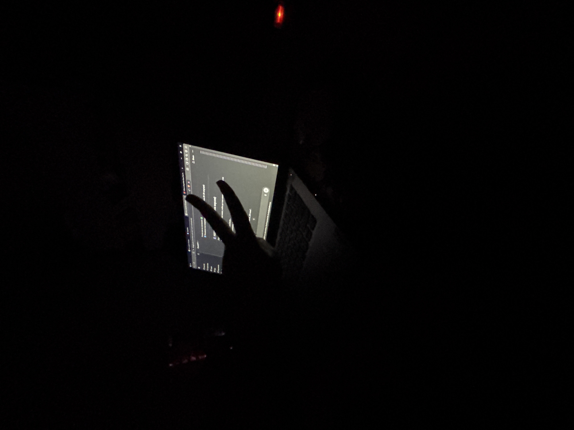 The image shows a dimly lit room with a laptop displaying a web page. A hand is gesturing in front of the screen, forming a "peace" or "victory" sign. The laptop is resting on a surface that appears to be a keyboard. Above the laptop is a red light, possibly from an electronic device. The overall environment is dark. There is no evidence to support the image being in Dobrovnitsa, Bulgaria.