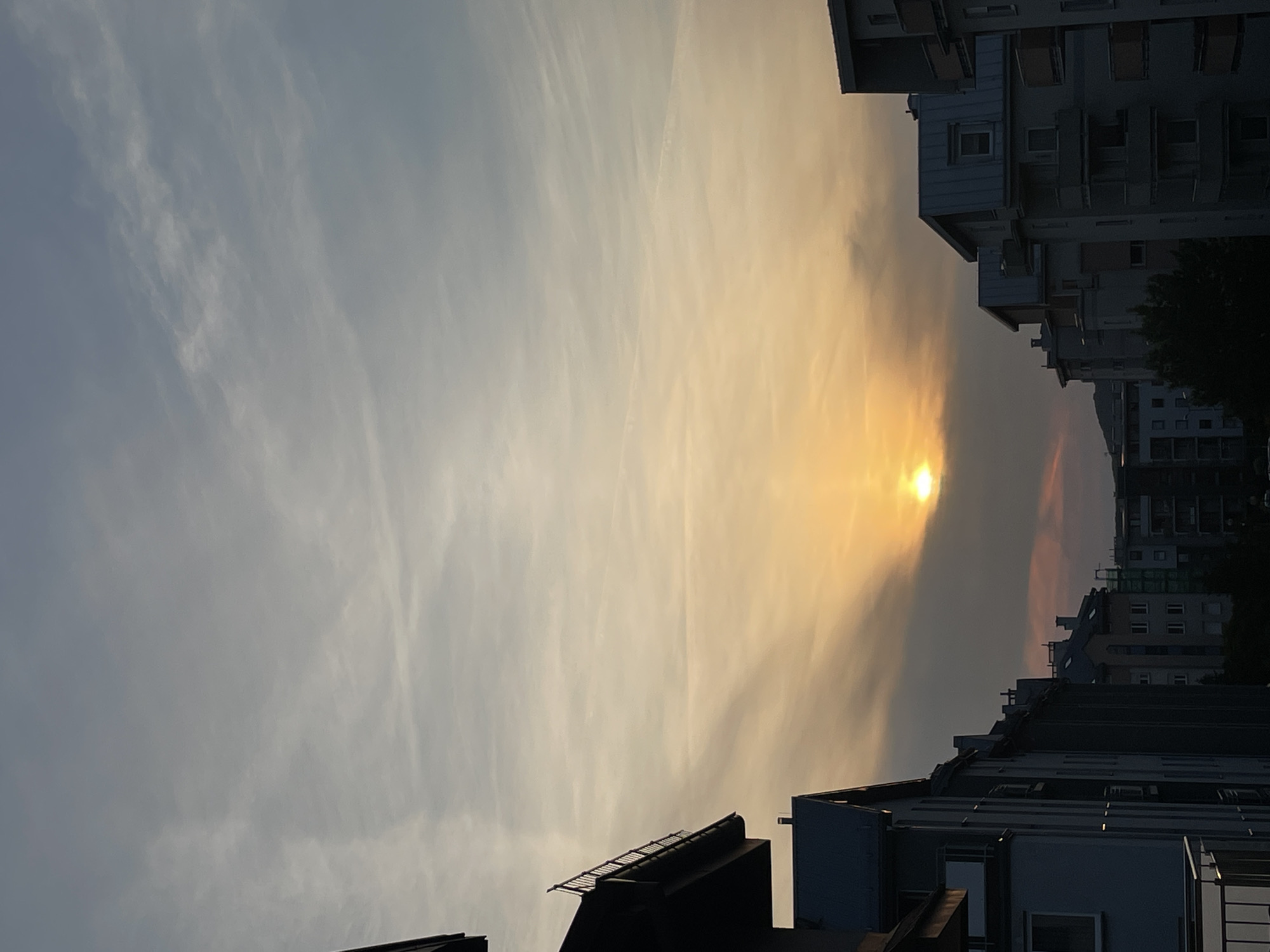 The image depicts an urban or suburban landscape captured during either sunrise or sunset. The sky occupies the majority of the frame, showcasing a gradient of colors from a pale blue-grey on the left to warm golden and orange hues towards the right. A bright, golden sun, partially obscured by clouds, is visible on the right side, casting a soft, luminous glow. Wispy, elongated clouds, possibly cirrus or altostratus, are stretched across the sky, catching the light and adding texture.
Silhouetted against the brightly lit sky are several multi-story buildings, likely residential or commercial structures. Their dark forms allow for the subtle recognition of architectural details such as balconies, numerous windows, and varied rooflines, some of which appear to have small structures or equipment. The buildings line the lower edge of the image, appearing densely packed on the right side and extending into the distance. There are no people or specific activities visible. The architectural style suggests a modern setting. No text is visible in the media file. The overall impression is one of serene beauty at the beginning or end of the day.