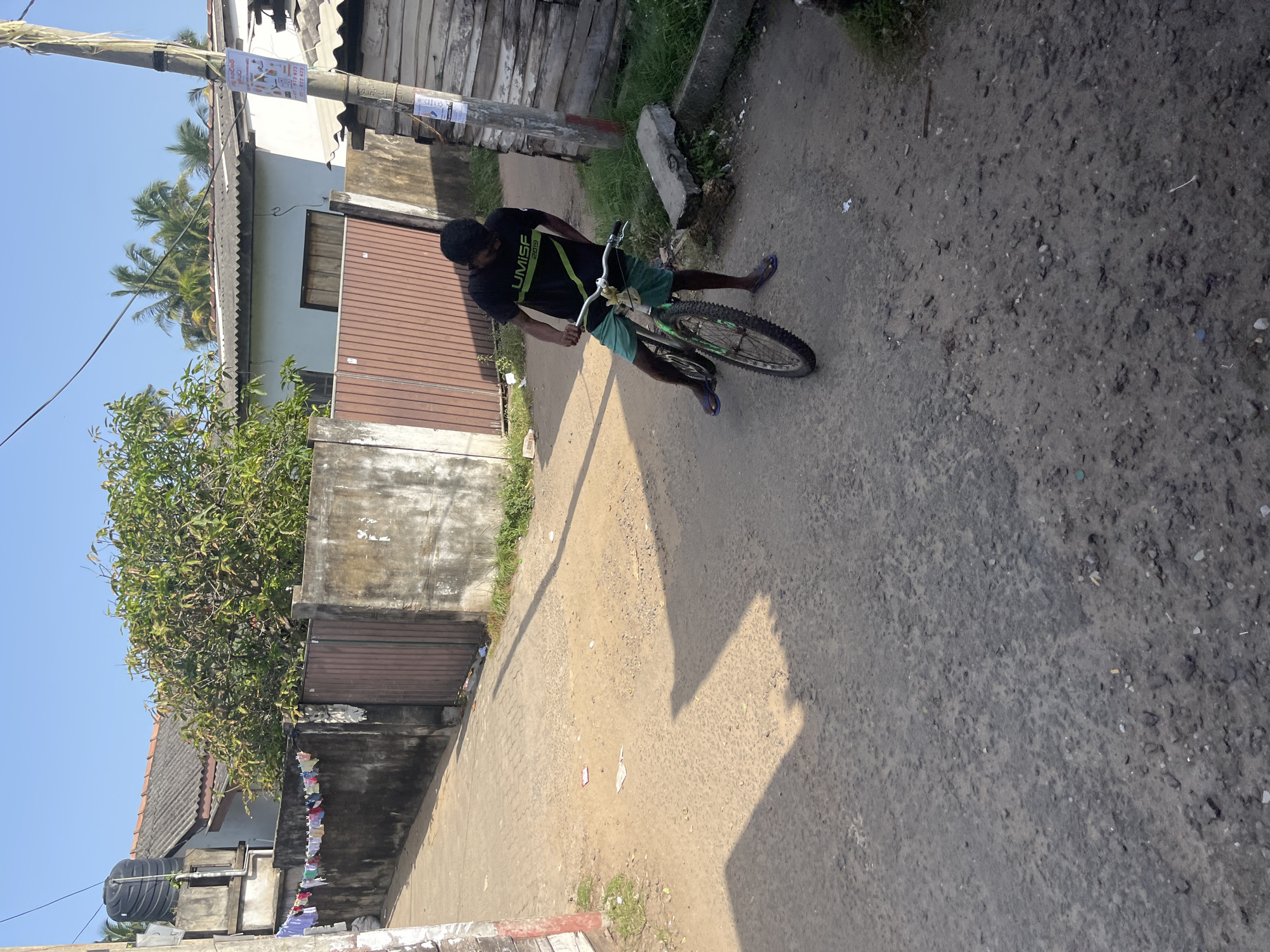 In Moratuwa, Sri Lanka, a young man in a black t-shirt and green shorts stands with a bicycle on a dusty, unpaved street. The bicycle is a mountain bike with a green frame and a white handlebar. The man is looking down at the bicycle. The street is lined with a weathered concrete wall on the left, on which laundry is hanging, and a wooden fence on the right. Behind the wall and fence, there are houses with tiled roofs and trees. The sky is clear and blue. The scene is illuminated by bright sunlight, casting shadows on the ground.