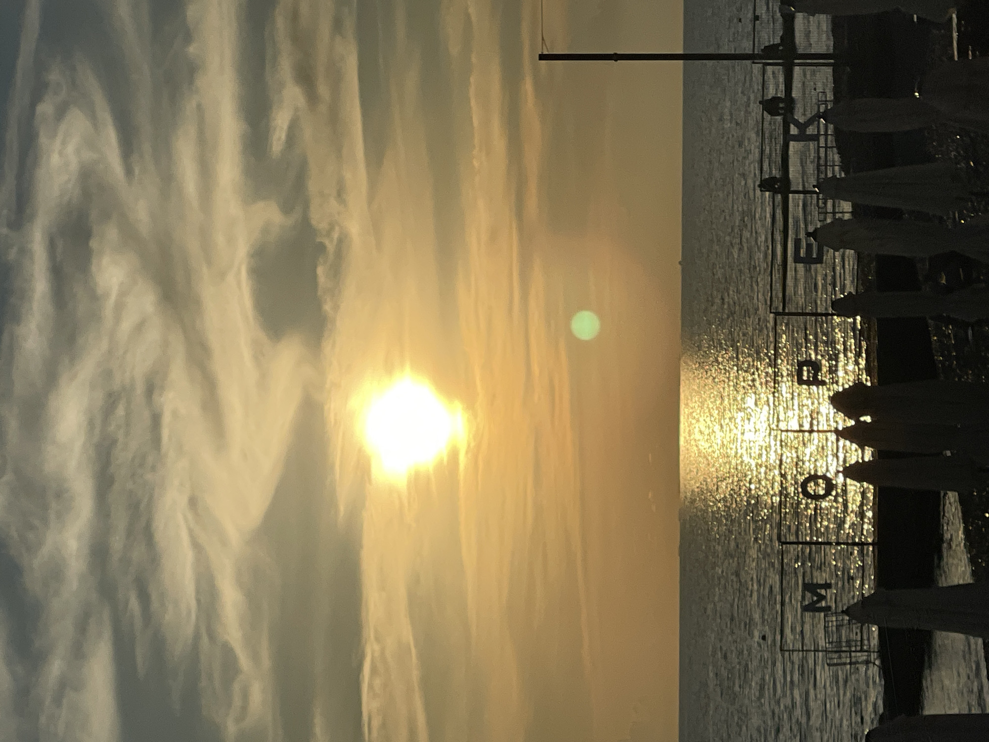 This is a landscape photograph taken at sunset on the coast of Sochi, Russia. The sun is a bright, white orb in the upper center of the image, casting a golden reflection on the calm sea. The sky is filled with wispy clouds, tinged with orange and yellow hues from the setting sun. In the foreground, a dark silhouette of beach umbrellas lines the shore. Behind them, a metal fence is visible with letters that appear to spell "MOPE". Several figures can be seen sitting on benches behind the fence. The sea stretches to the horizon, where distant ships are faintly visible.