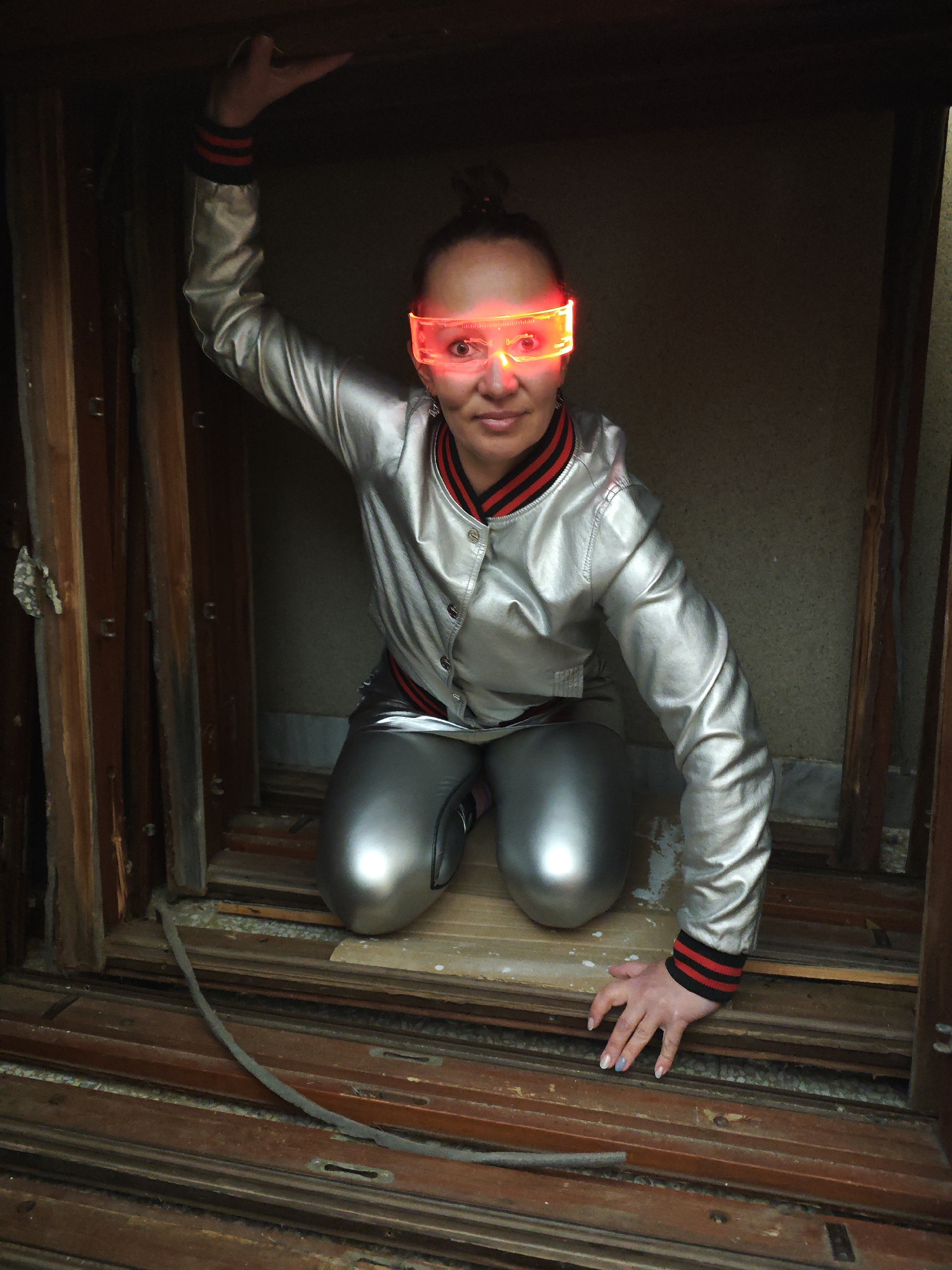 A woman is kneeling in a confined, weathered wooden structure, possibly an abandoned window frame, in what appears to be an indoor or enclosed space. The location is Марковский, Russia. She is wearing reflective silver pants and a matching jacket with red and black striped trim on the cuffs and collar. The woman also sports light-up red glasses. Her hair is pulled back into a bun. Her left hand is raised towards the top of the frame, while her right hand rests on the wooden surface in front of her. The overall scene has a somewhat gritty, urban decay aesthetic.