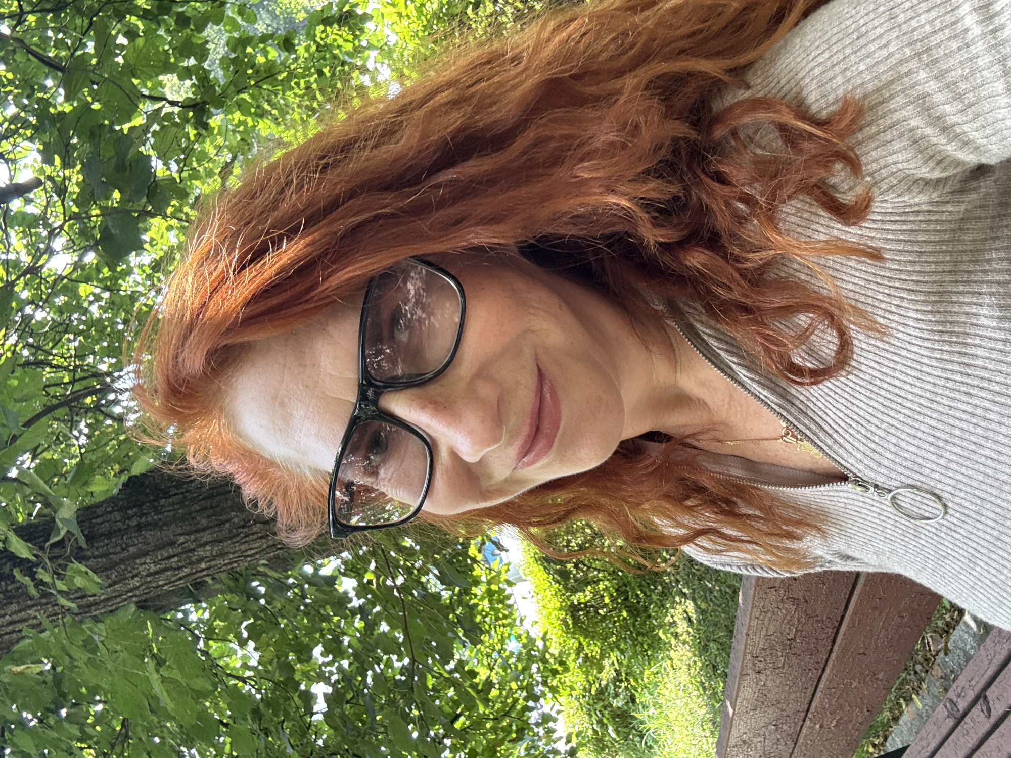 A woman with long, wavy, ginger hair and fair skin, wearing glasses and a grey ribbed sweater, is pictured outdoors. She is sitting on a wooden bench, with a tree and greenery in the background. The scene appears to be a park in Zelenograd, Russia.