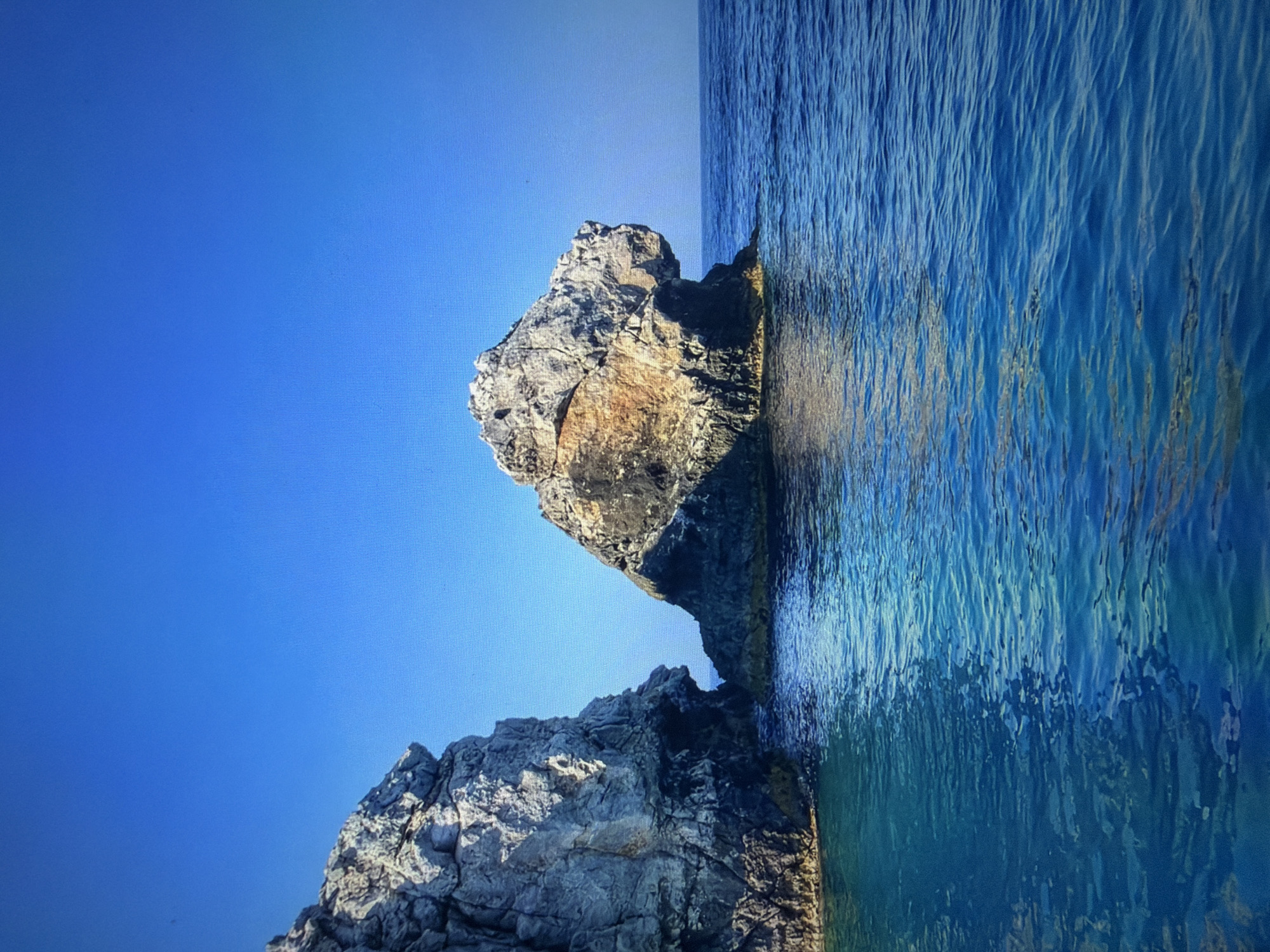 A rocky outcrop stands in bright blue ocean water under a clear, deep blue sky. The rocks are textured and appear weathered, with some areas showing brown and white coloration, contrasting with darker shaded areas. The water is calm with gentle ripples reflecting the sunlight. The scene suggests a natural coastal environment, possibly a remote island or a rugged coastline. The image is a landscape photograph. There is no indication of any specific city, including Kotelniki, Russia, or any people, objects, actions, or interactions.
