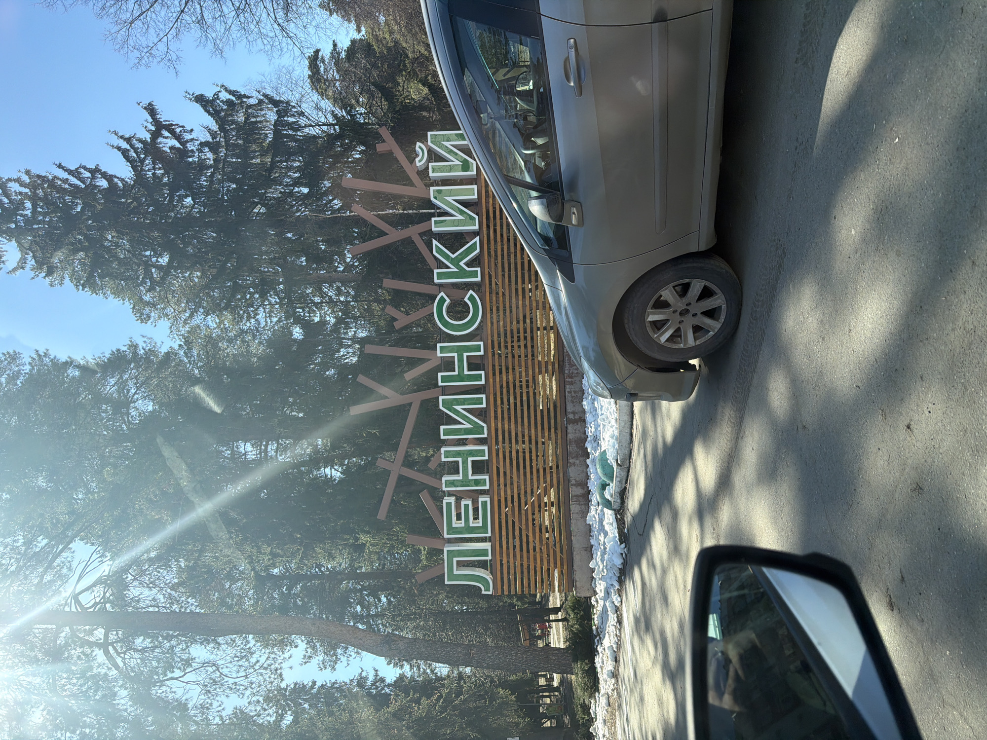 The image shows a park entrance sign with the word "Ленинский" (Leninsky) in Cyrillic. The sign is made of wooden planks and brown wooden rods shaped like branches. The letters are green with a white border. A silver car is parked next to the sign, partially obscuring it. Trees are visible in the background. The photo appears to have been taken from a car, with a side mirror visible in the lower right corner. Snow patches are on the ground, suggesting a cold season. The location is likely Ленинский (Leninsky) Park in the city of Sarapul, Russia, based on the sign. The photo was taken during daylight hours, likely in a sunny environment.