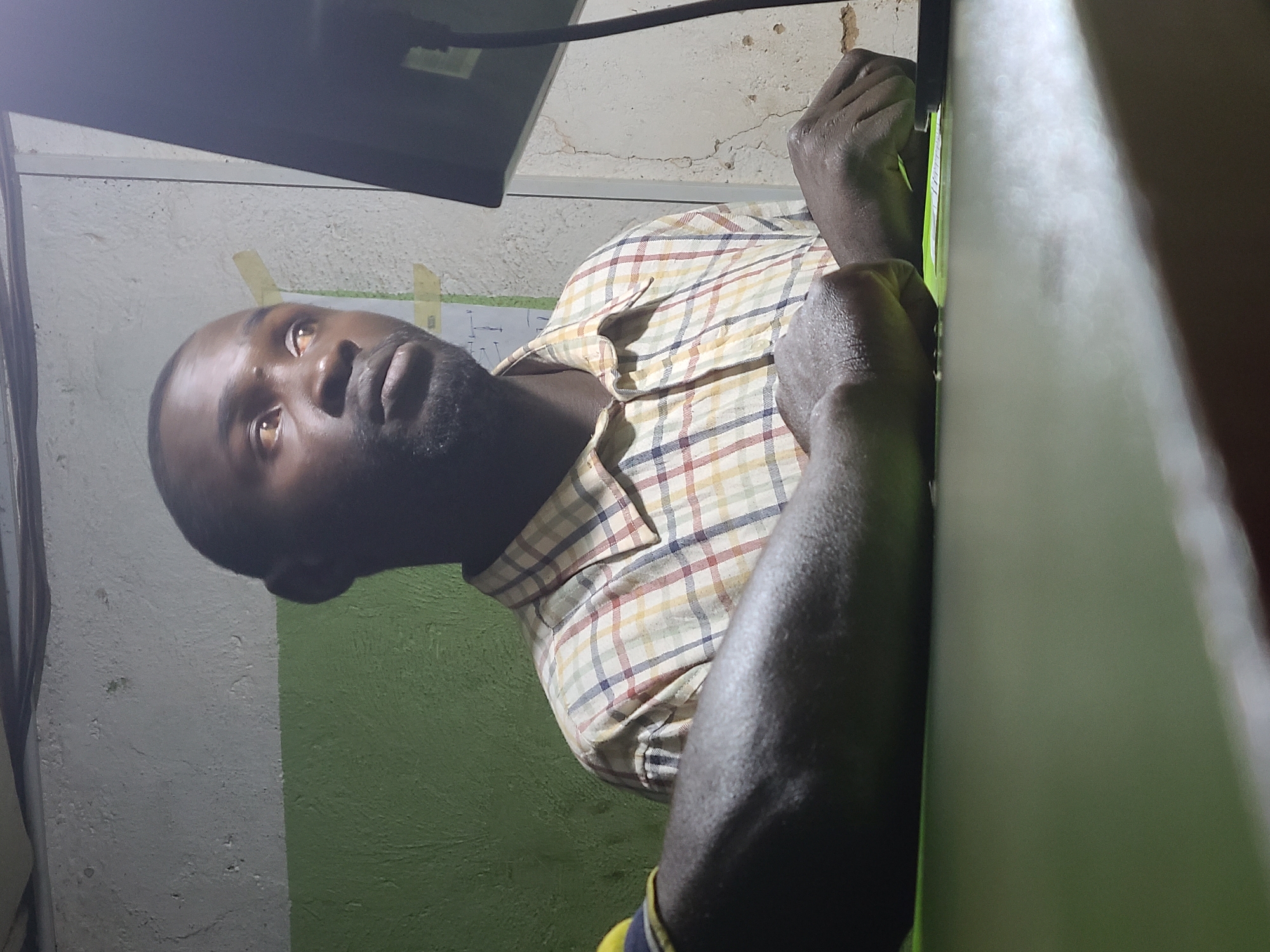 A dark-skinned man with short hair and a beard is indoors, wearing a plaid shirt. His arms are visible, with one hand gripping a green object or surface. He appears to be looking upward towards an unseen object, possibly a monitor or screen partially visible at the top of the frame. The background shows a wall that is painted white and green, and there are pieces of tape on it. The location is identified as Jos, Nigeria.