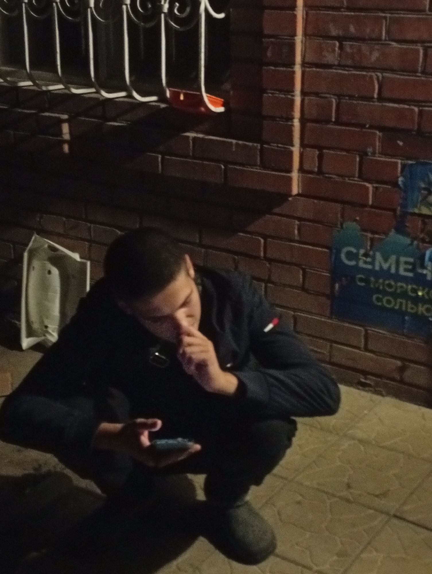 The media file shows a young man crouched on a tiled surface, looking at his phone. He is wearing a dark jacket and pants. The man's face is partially obscured by his hand, and he appears to be deep in thought or concentrating on his device.
The setting appears to be outdoors at night, with artificial lighting casting shadows on the brick wall behind him and the ground in front. To the left of the man, a white plastic object, possibly a piece of discarded furniture or a large container, is leaning against the wall. The brick wall has a weathered texture, and a portion of a poster or advertisement is visible on it, featuring blue text that reads "СЕМЕ" and "С МОРСКО" and "СОЛЬЮ". The iron bars of a window or railing are visible at the top of the frame.
The overall mood is quiet and solitary, with the focus on the man and his interaction with his phone in a dimly lit environment. The time of day is clearly night, and the weather is not discernible. The location context provided is "Маяк, Russia."
