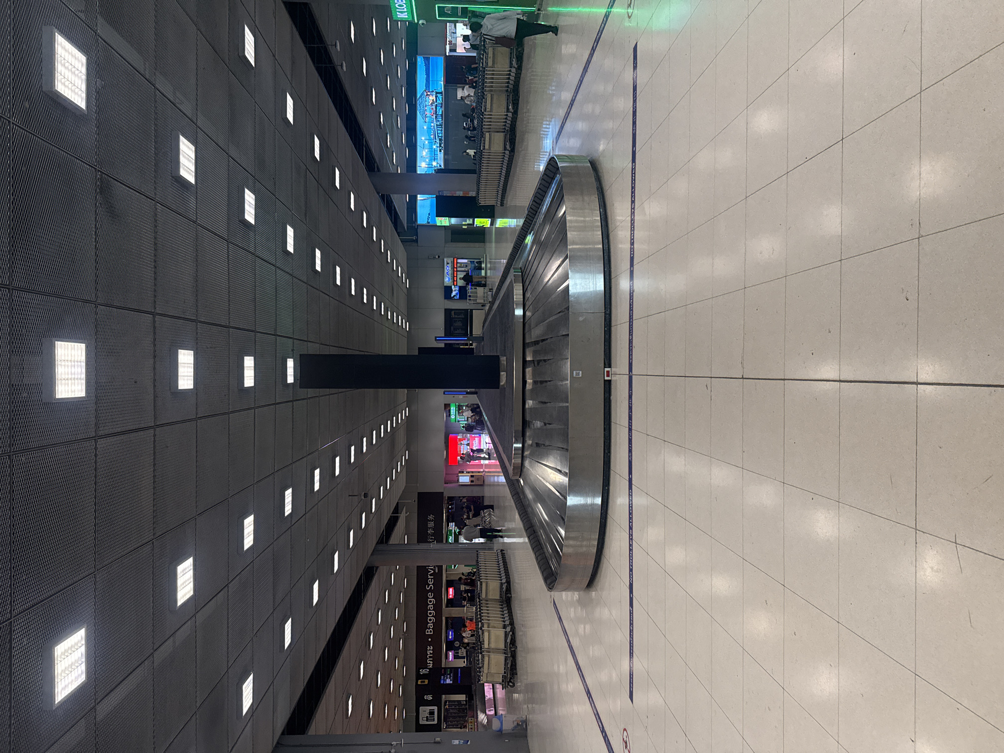 The image shows an empty baggage claim area in an airport terminal. A large, circular baggage carousel is in the foreground, with a black pillar behind it. The floor is tiled with light-colored squares, and blue lines with text are present on the floor, indicating a walkway or restricted area. In the background, several rows of luggage carts are parked. There are also signs and screens visible, including one with the text "Baggage Servi" and another displaying a scenic image of a pier. A few people are scattered throughout the area, some standing or walking. The ceiling is a grid of acoustic tiles with rows of fluorescent lights. The scene appears to be in an indoor, public space, likely an airport due to the presence of the baggage carousel and luggage carts. The location is specified as Nong Prue Subdistrict, Thailand.