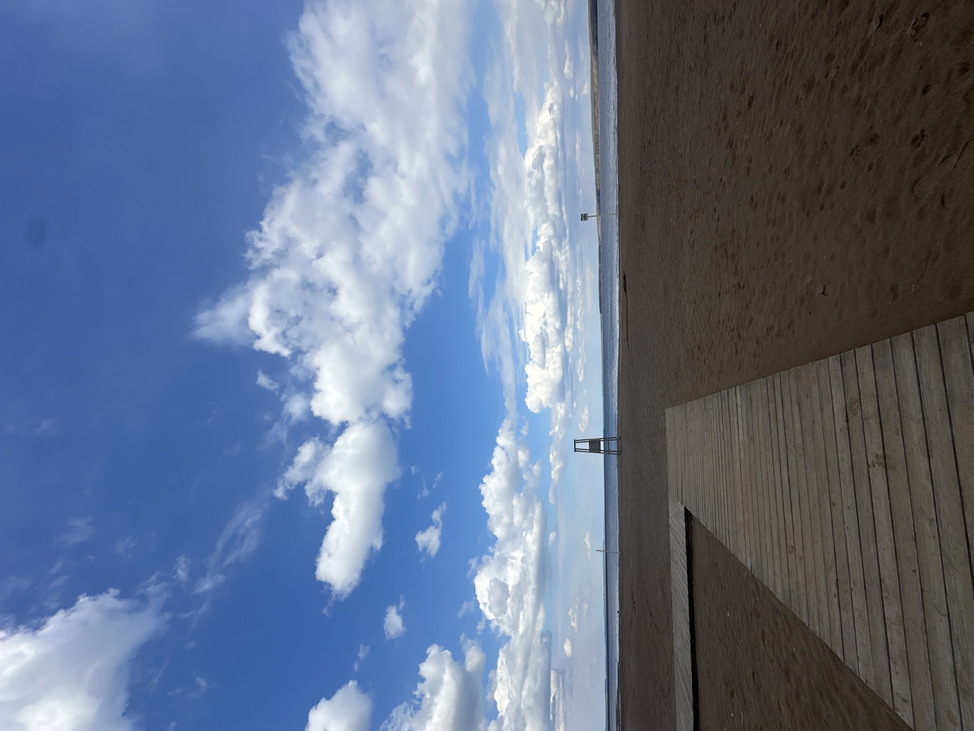 The image shows a beach scene in the city of Chaikovsky, Russia. The sky is blue with scattered white clouds. The beach is sandy and there is a wooden boardwalk extending into the sand. In the distance, the horizon line of the water meets the sky. There is a lifeguard stand visible on the beach.