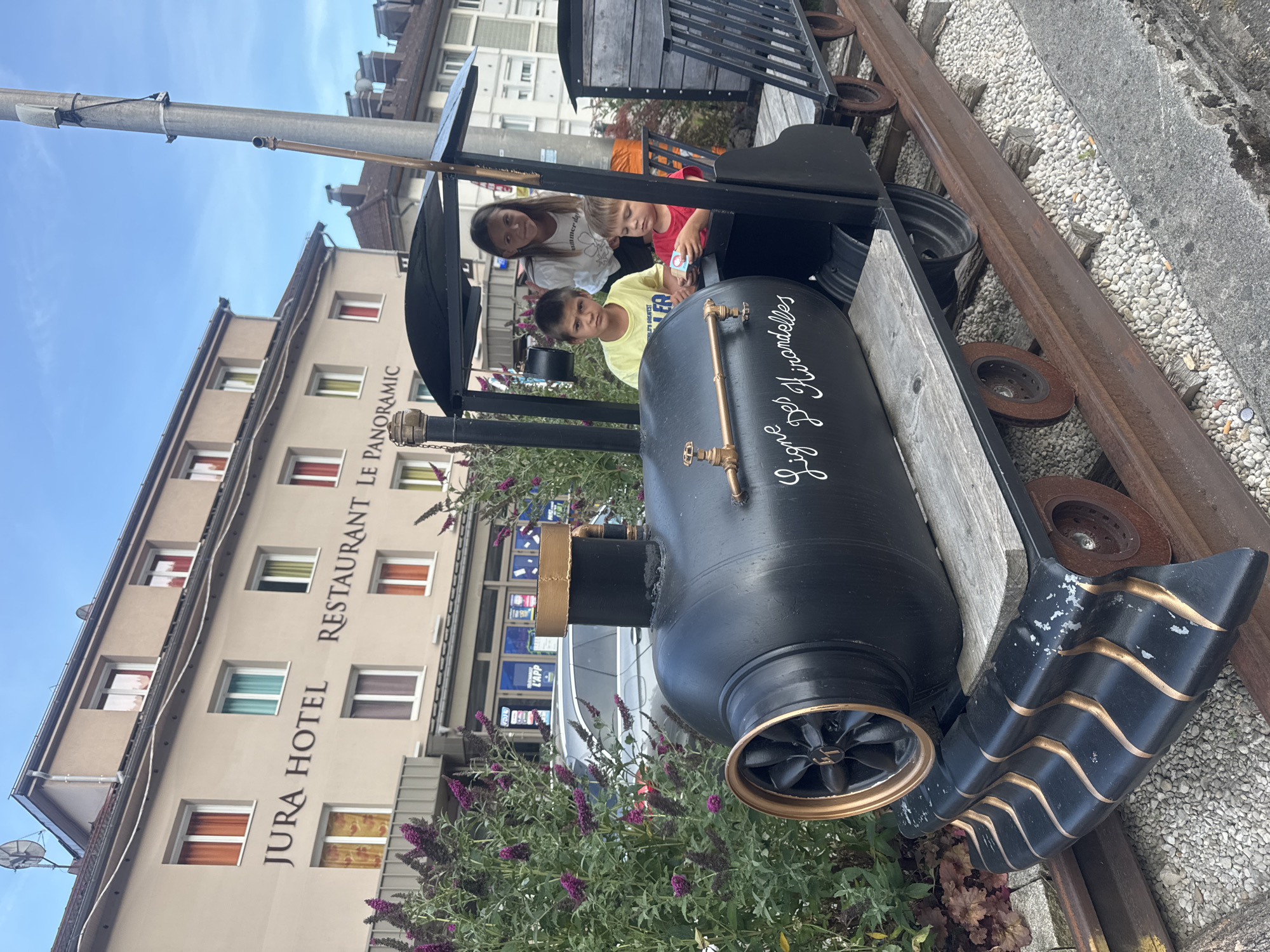 In Saint-Claude, France, in front of the Jura Hotel and Restaurant Le Panoramic, two young boys and a young girl are sitting on a miniature train. The train is black with gold accents and has "Ligne Des Hirondelles" written on its side in white cursive. The older boy wears a yellow t-shirt, the younger boy a red t-shirt, and the girl a white t-shirt. The train sits on a track with gravel on either side. Purple flowers are visible to the left of the train.