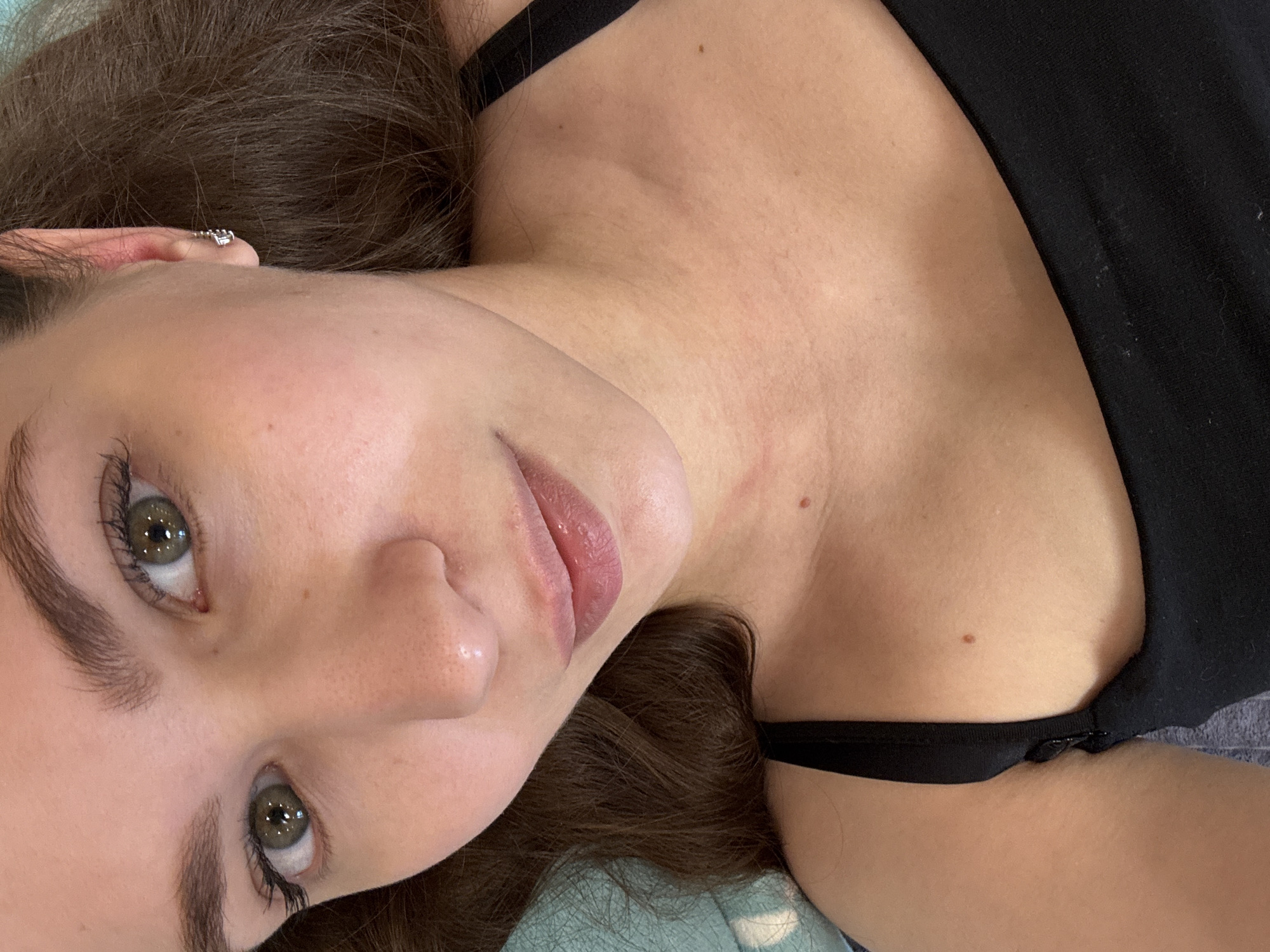 A close-up, angled portrait captures a young woman looking directly at the viewer. She is positioned on her back, with her head turned to her right, and her dark brown hair spread out around her. Her face is in the foreground, featuring clear, green-hazel eyes with visible eyelashes, and full lips that are slightly parted. A small, silver-colored earring with multiple facets or stones is visible in her earlobe.
Her skin has a clear complexion, with a few small moles visible on her neck and upper chest. She is wearing a black strap top, the fabric of which is visible around her shoulder and neck. The background shows a light blue, textured fabric, suggesting she is lying on a soft surface, possibly a bed or couch, indoors. The lighting is soft and even, without harsh shadows, indicating an indoor setting. The image evokes a calm or contemplative mood. No text is visible.