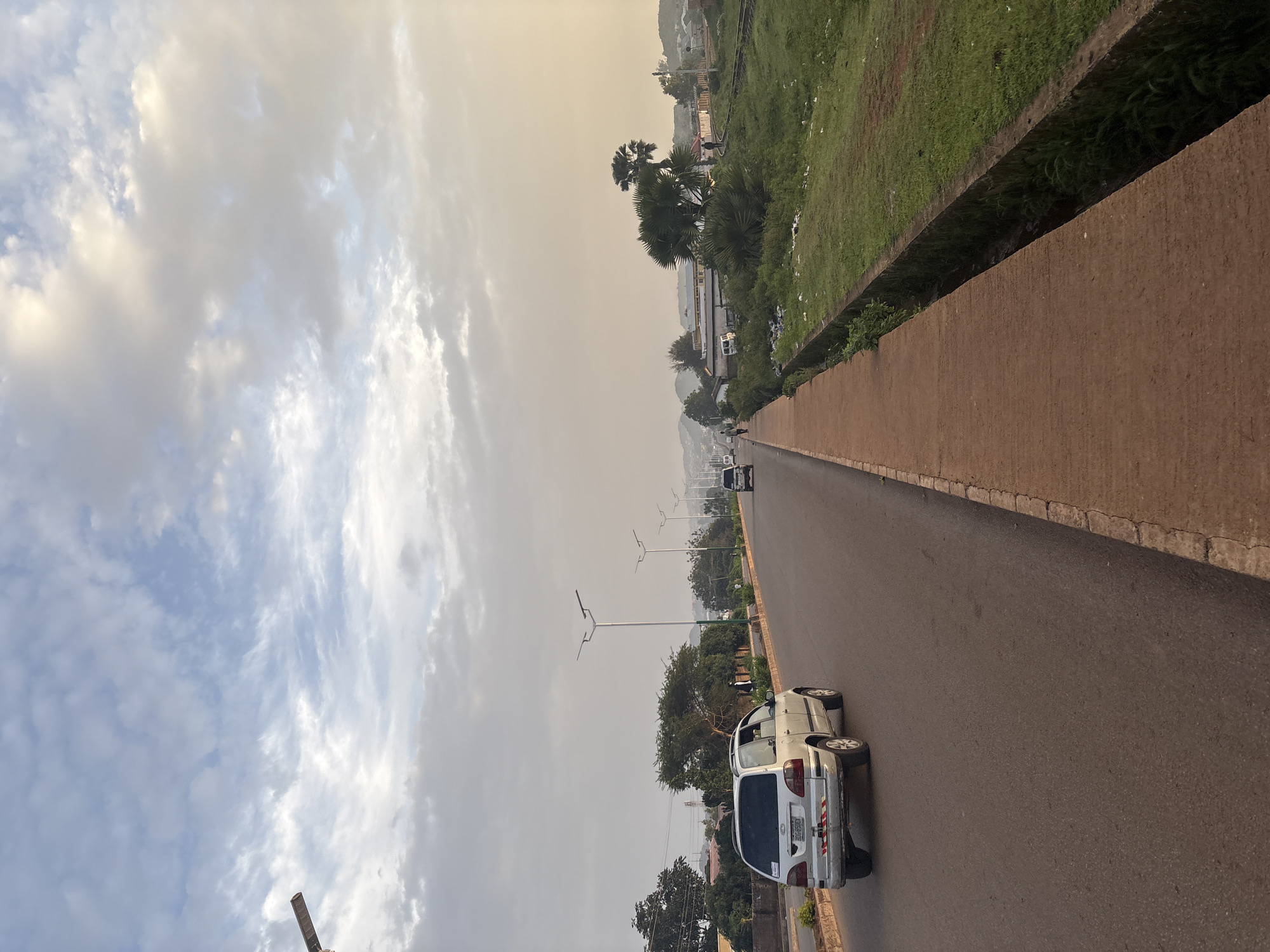 The image depicts a road in Bukuru, Nigeria, under a cloudy sky. A silver minivan is driving on the left lane of the asphalt road, facing away from the camera. A sidewalk made of red earth runs along the right side of the road, bordering a grassy embankment. In the distance, several streetlights line the road, and more vehicles are visible. Palm trees and other vegetation are scattered along the sides of the road, with buildings and hills in the background. The scene appears to be during daytime, possibly late afternoon, given the soft lighting.