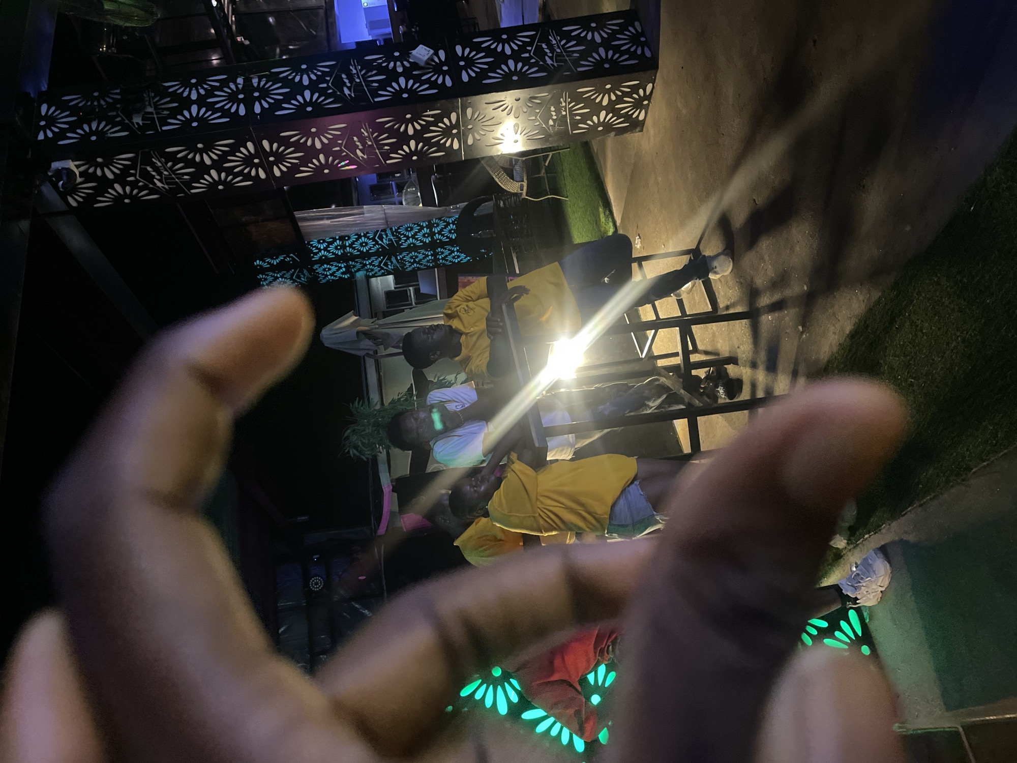 A person's blurred hand is in the foreground, obscuring the lower left portion of the image. Behind the hand, four young adults, two males and two females, are gathered in an outdoor setting at night. One male, wearing a white t-shirt and jeans, is leaning on a railing and looking to the right. Another male, wearing a yellow polo shirt and dark pants, is leaning on a metal chair and looking to the left. A female wearing a yellow t-shirt and denim shorts is standing and talking to someone off-camera. Another female, wearing a red top and dark pants, is partially visible in the background. The scene is illuminated by green neon lights shaped like petals on a dark wall to the left and by a bright light source emanating from the center right of the image, casting long shadows. Decorative metal columns with intricate cutouts are visible on the right side of the image, and a dark ceiling structure is above. The ground appears to be a mix of concrete and artificial grass.