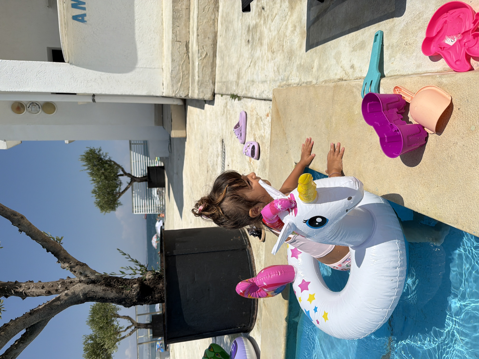 A young girl is playing in a swimming pool with a unicorn-shaped inflatable ring. She is wearing a pink swimsuit and has her hair pulled back. The pool is clear blue and reflects the sunlight. Several beach toys, including a purple bucket and a blue plastic rake, are scattered on the concrete poolside. The scene suggests a warm, sunny day, likely in the afternoon, given the bright lighting and shadows. In the background, there are trees and a view of the sea, with people and umbrellas visible on a beach, suggesting a coastal location. The overall mood appears playful and carefree. There is some visible text on the white structure in the upper left corner, which appears to be "AM". The context provided indicates the location is Burgas, Bulgaria.