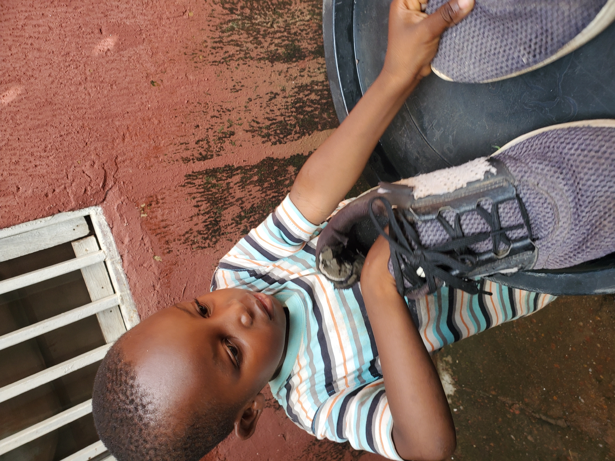 This media file shows a young boy from the side, looking up and to the left. He is wearing a horizontally striped t-shirt with blue, orange, and white stripes. His hair is short and dark. He is holding a dark-colored sneaker with black laces. The sneaker appears to be a bit worn. His right arm is visible, and he is holding the sneaker with his hand.
The background is a reddish-brown surface, possibly a wall or pavement, with some darker patches that might be moss or dirt. To the left of the boy, there is a white slatted structure, resembling a grate or vent. The lighting suggests it is daytime, with some shadows present. The overall scene appears to be outdoors, possibly in a residential area in Jos, Nigeria. The boy's expression is neutral, and he seems to be focused on the shoe. The image is oriented sideways.