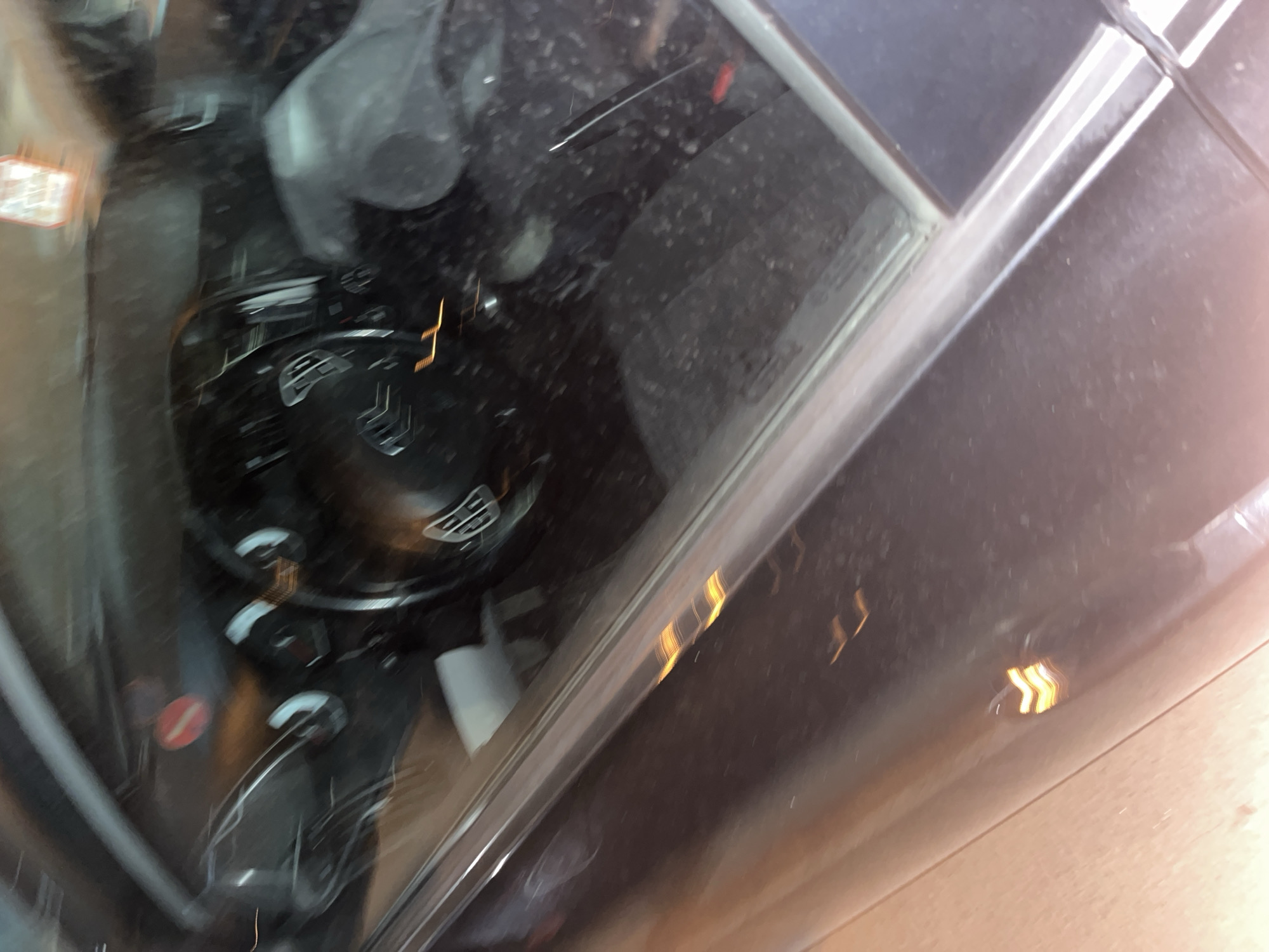 View from inside a car looking towards the passenger side. The dashboard and steering wheel are visible through the windshield. The car is likely parked, judging by the angle and reflection. The exterior of the car is visible on the right side of the frame, and a blurred image of an urban environment can be seen outside the windshield. Given the prompt's location constraints, the urban setting is likely in Blagoevgrad, Bulgaria.